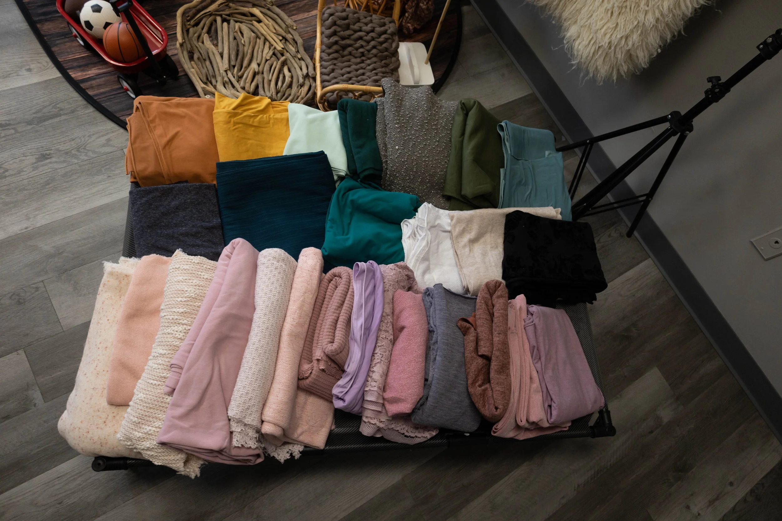 A black rolling cart filled with folded clothes of various colors, including pinks, grays, greens, blues, and neutrals, with a table in the background holding baskets and sports balls.