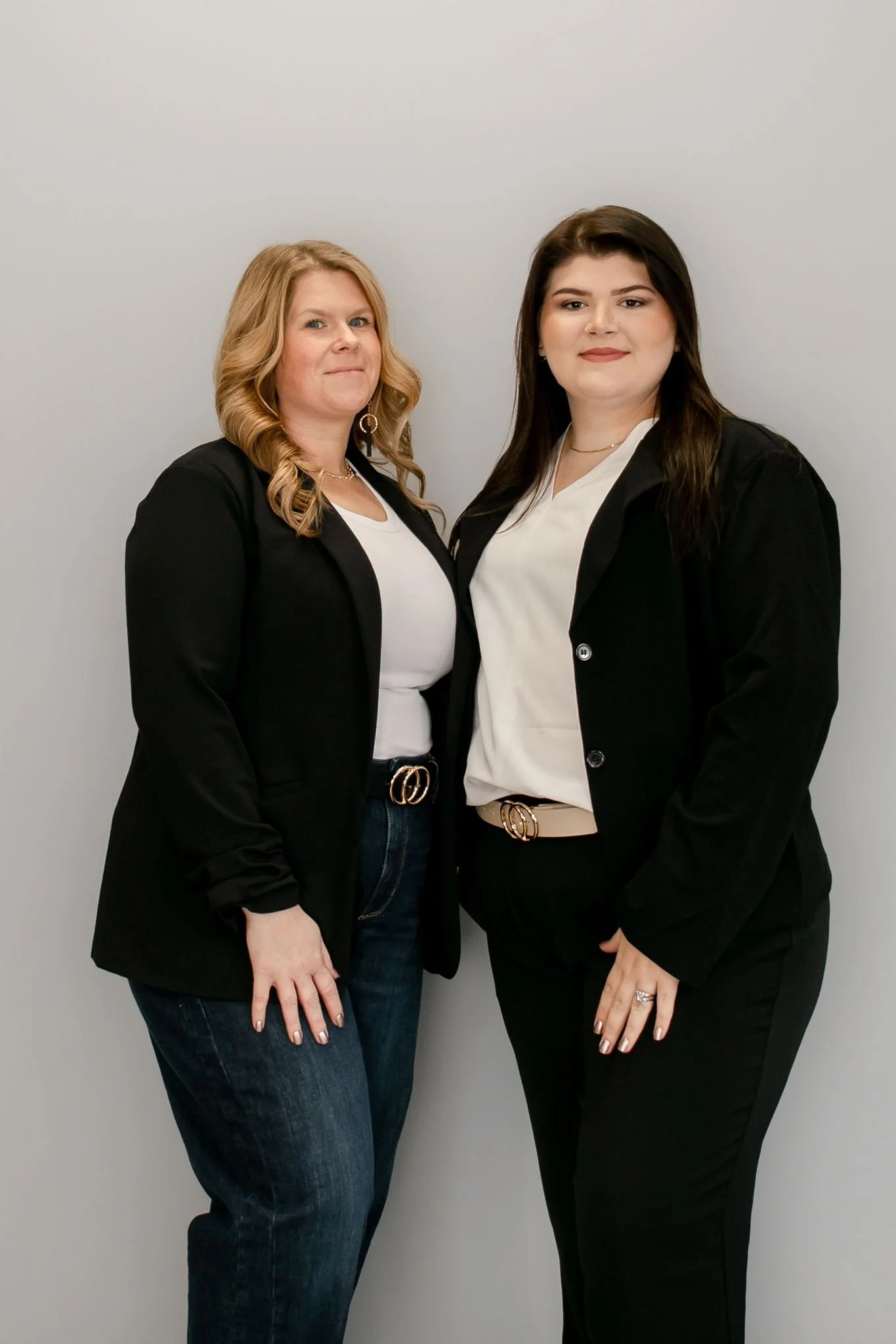 Two women standing side by side against a plain light-colored wall, wearing business casual attire.