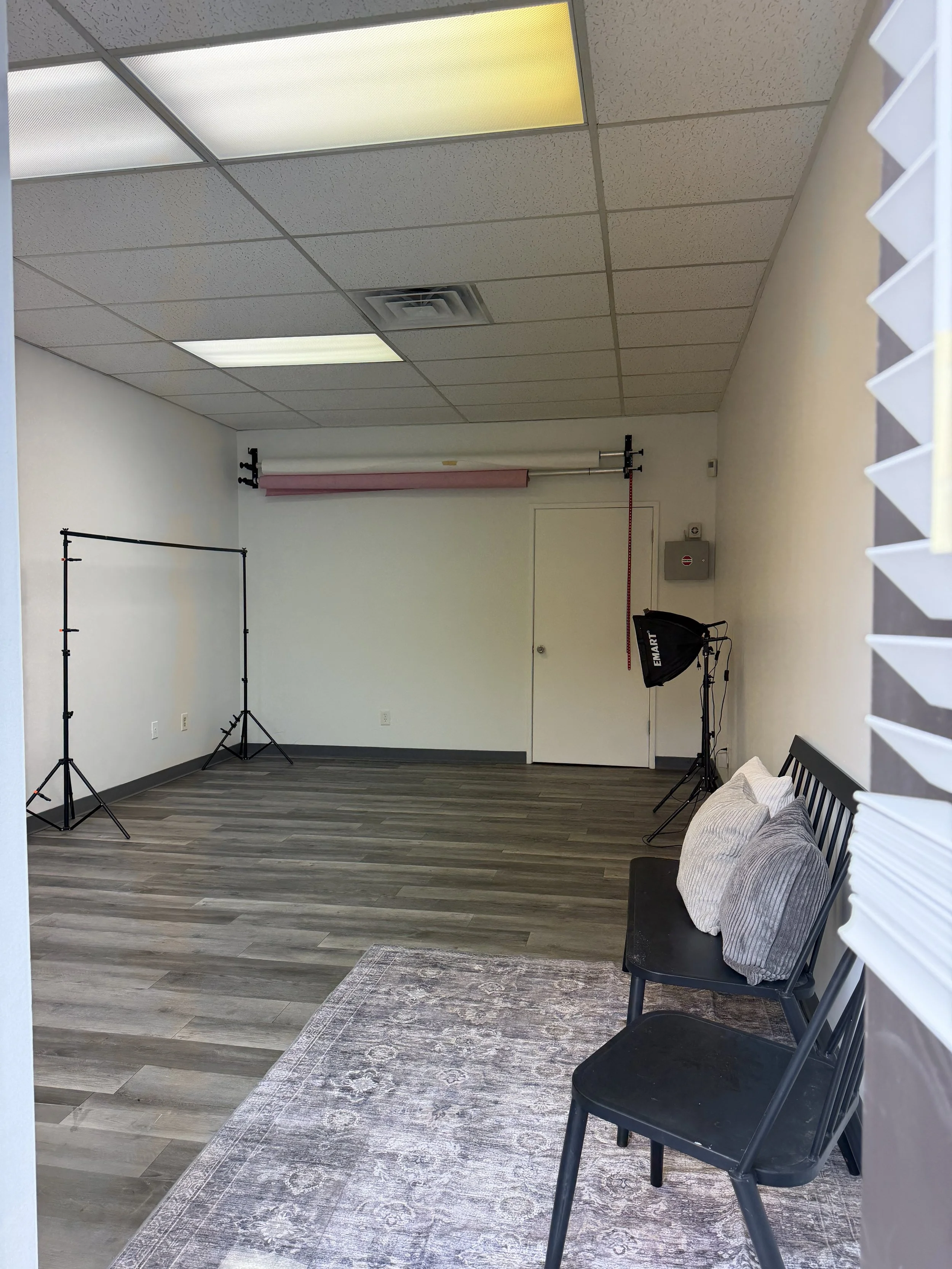 Photography studio with backdrop stand, studio lights, gray chairs on a rug, and ceiling fluorescent lights.