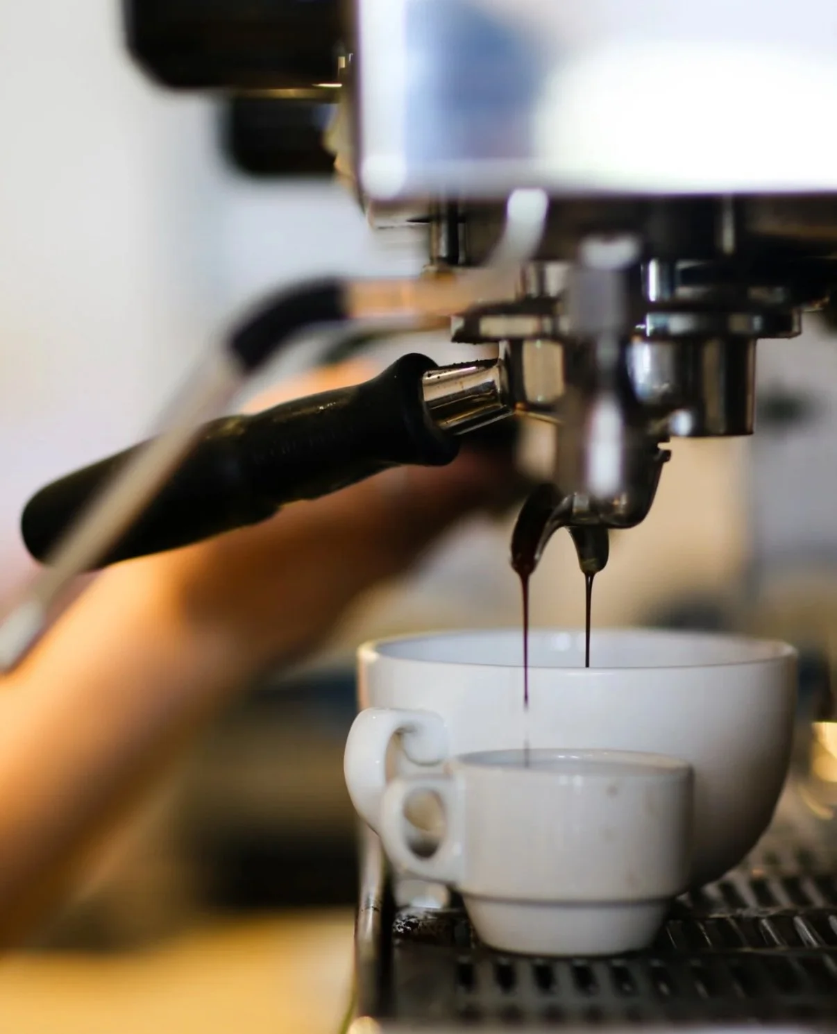 Image of espresso machine dripping coffee into two differently sized mugs with hand in the background.