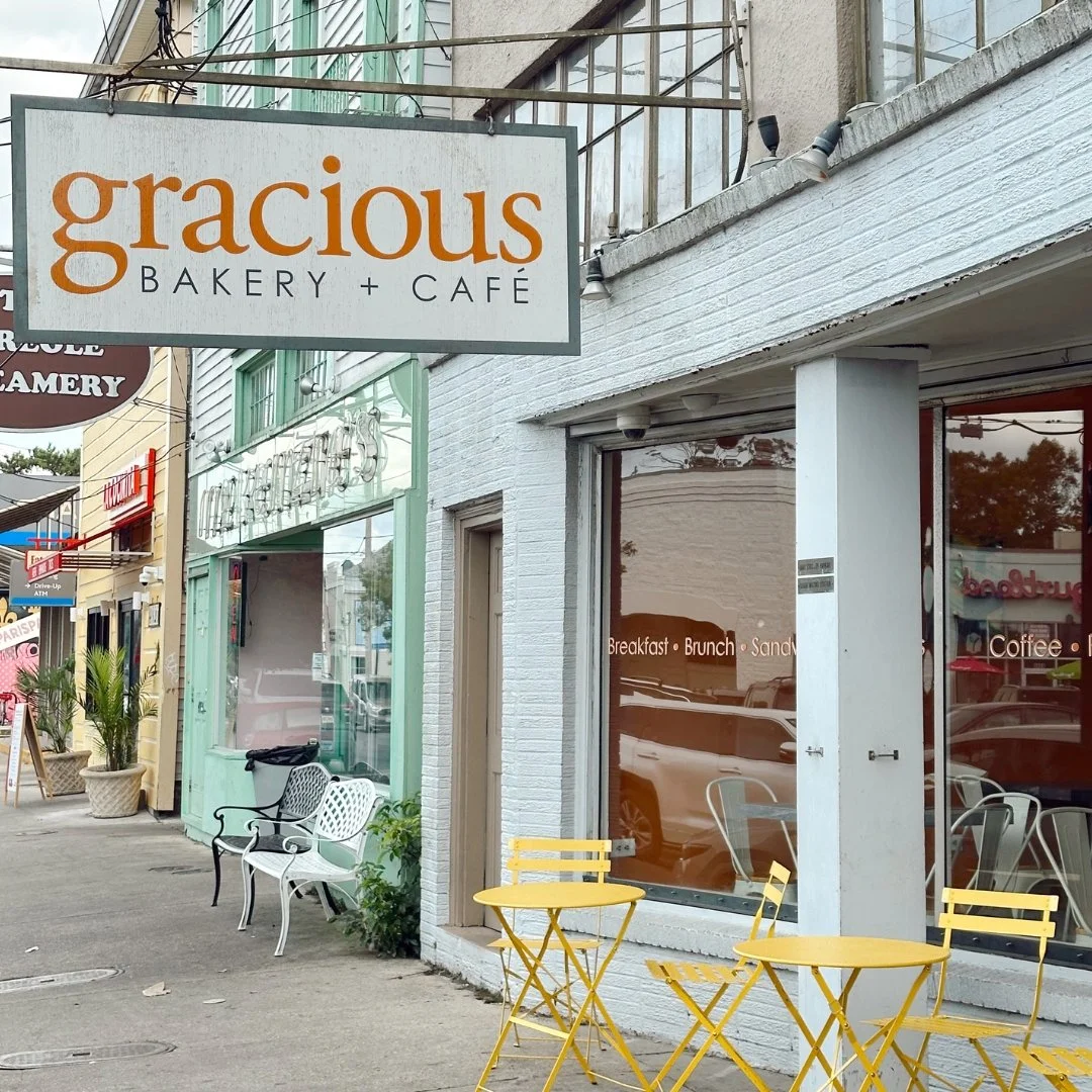 Street view of a bakery and cafe named "gracious" with outdoor seating including yellow tables and chairs, and white and black benches. The shop has large windows and signs for breakfast, brunch, sandwiches, and coffee.
