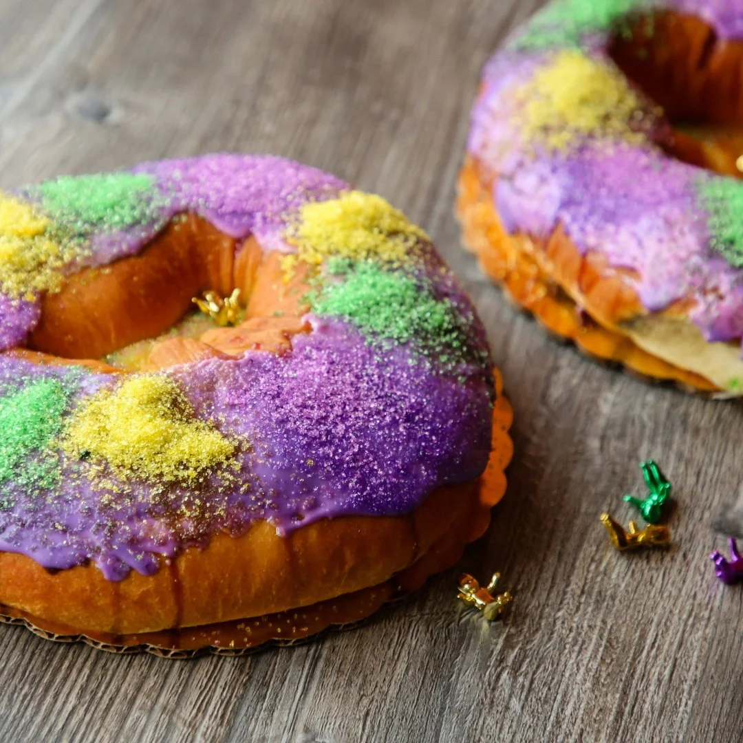 Two Mardi Gras themed donuts with purple, green, and yellow icing and sprinkles, resting on a wooden surface.