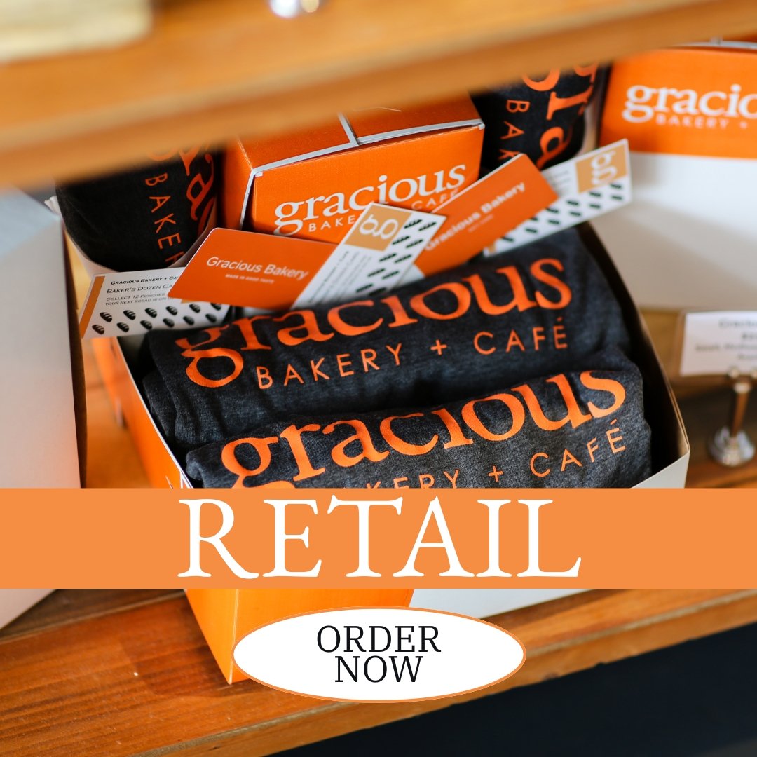 Retail display of Gracious Bakery + Café products, including boxes and towels with the bakery's logo, on a wooden shelf.