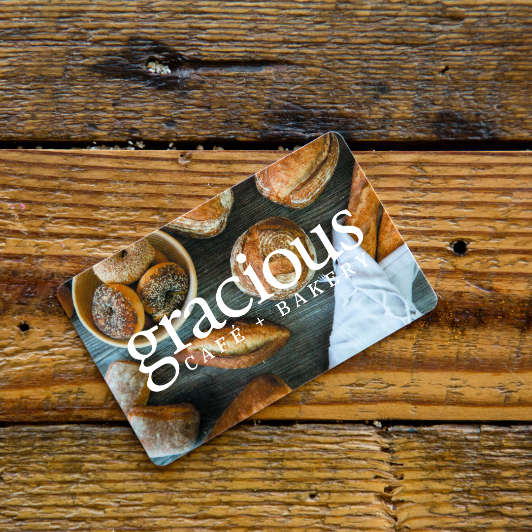 A business card for Graacious Cafe + Bakery on a wooden surface, featuring images of baked goods such as bagels and bread.