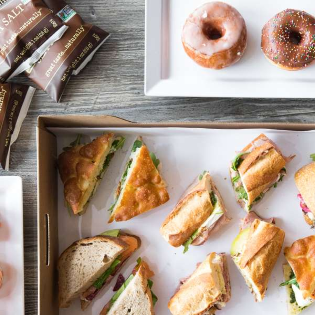 Assorted small sandwiches with meats, cheeses, and greens on a tray, two donuts with icing and sprinkles on a plate, and packets of salt on a wooden table.
