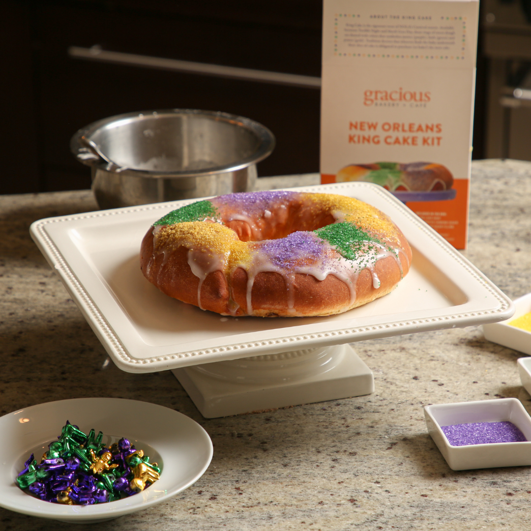 A Mardi Gras King Cake with white icing and colorful purple, green, and gold sprinkles on a white cake stand, with Mardi Gras beads in a white dish and a box of Gracious Gourmet Cake Kit in the background.