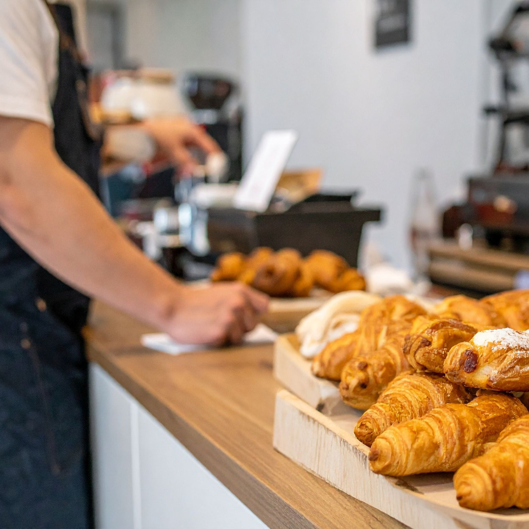 Display of assorted croissants and pastries on a wooden counter at a bakery with a person in the background.