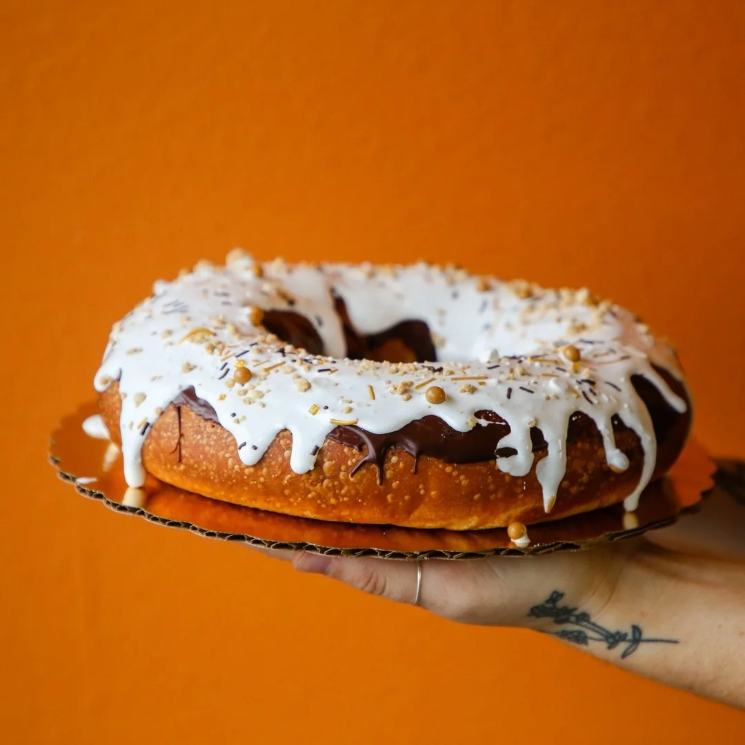 A hand holding a large donut with white, chocolate, and nut toppings against an orange background.