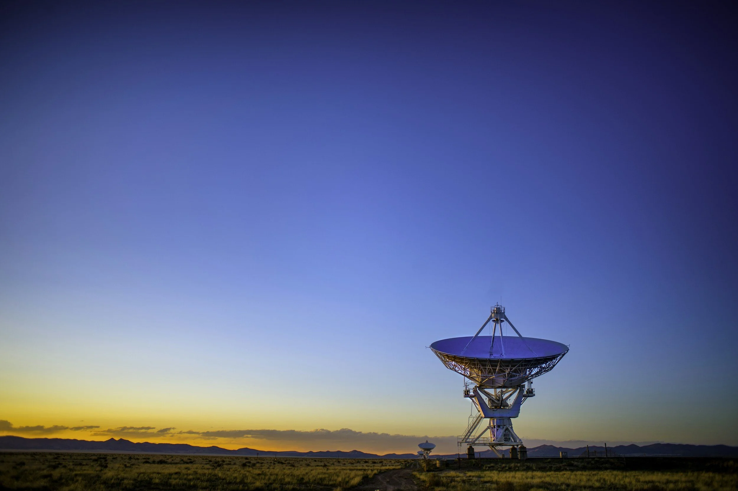 Large radio telescope dish in an open field during sunset with mountains in the background.
