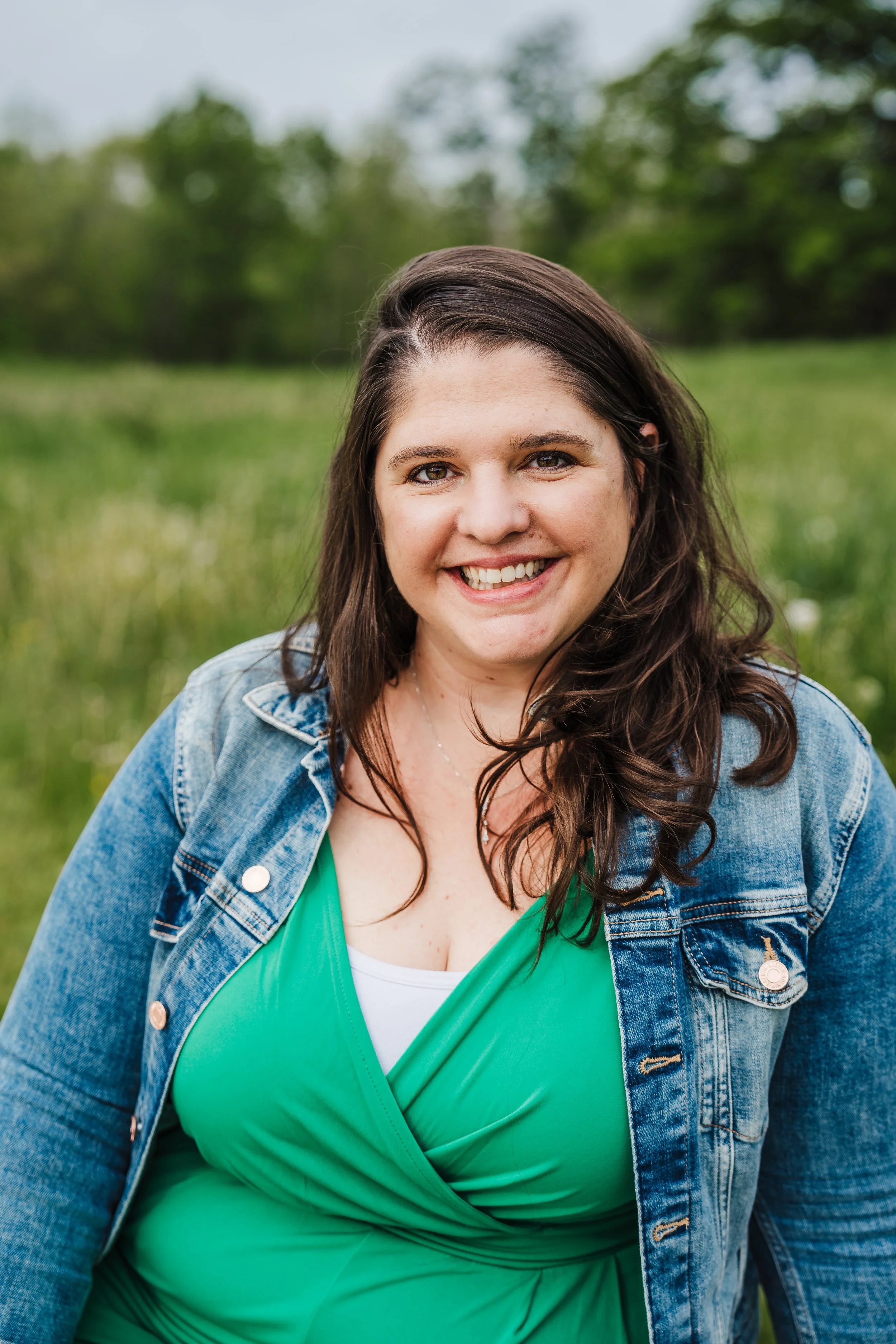A woman with long, dark brown hair, smiling and wearing a green top and a denim jacket, standing outdoors in a field of grass with trees in the background.