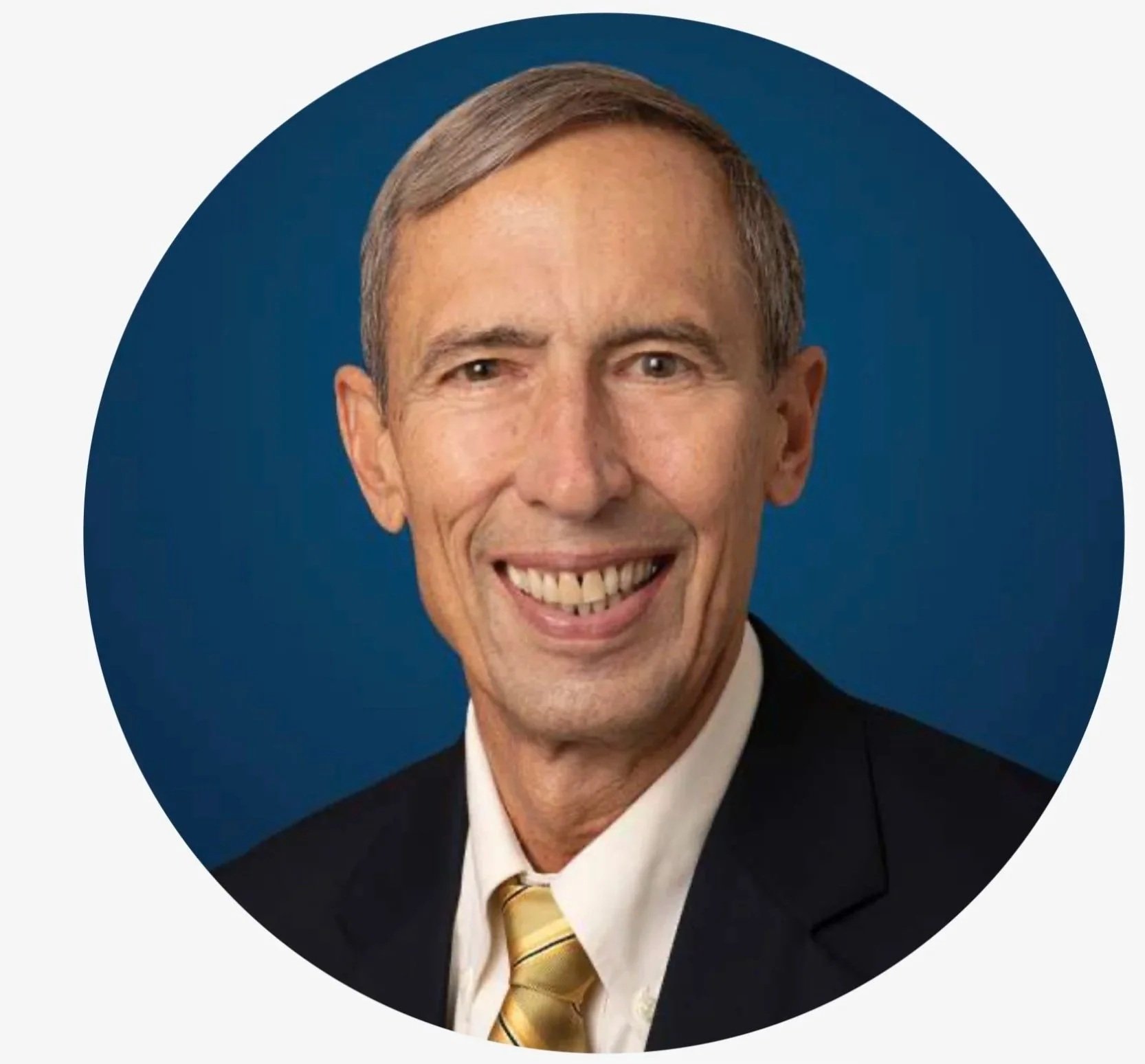 Headshot of a smiling man with gray hair, wearing a black suit, white shirt, and yellow striped tie, against a blue background.