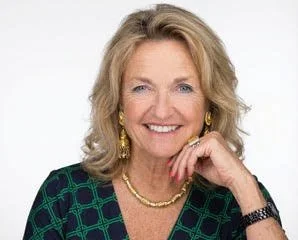 A woman with blonde hair, smiling, wearing a green patterned top, gold jewelry, and a watch, against a plain white background.