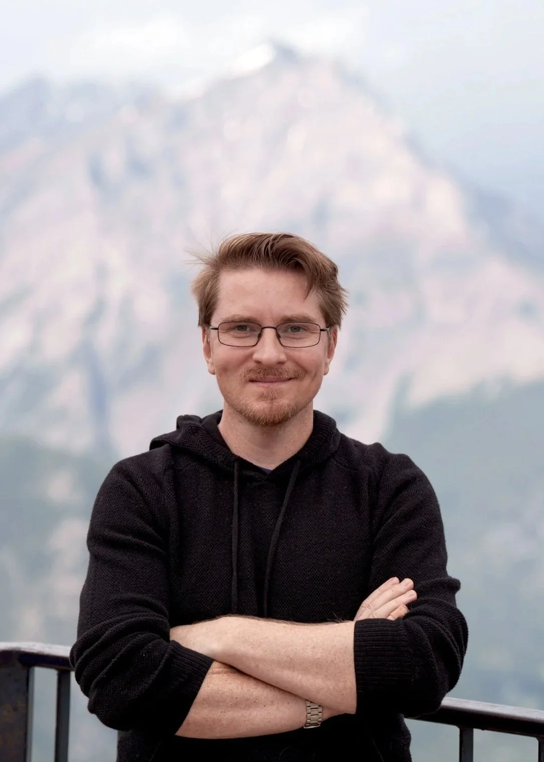 A man with glasses and a short beard, wearing a black hoodie, standing in front of a mountain.