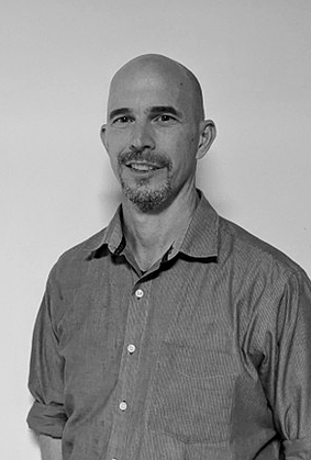 Black and white photo of a man with a bald head and goatee, wearing a collared shirt and smiling.