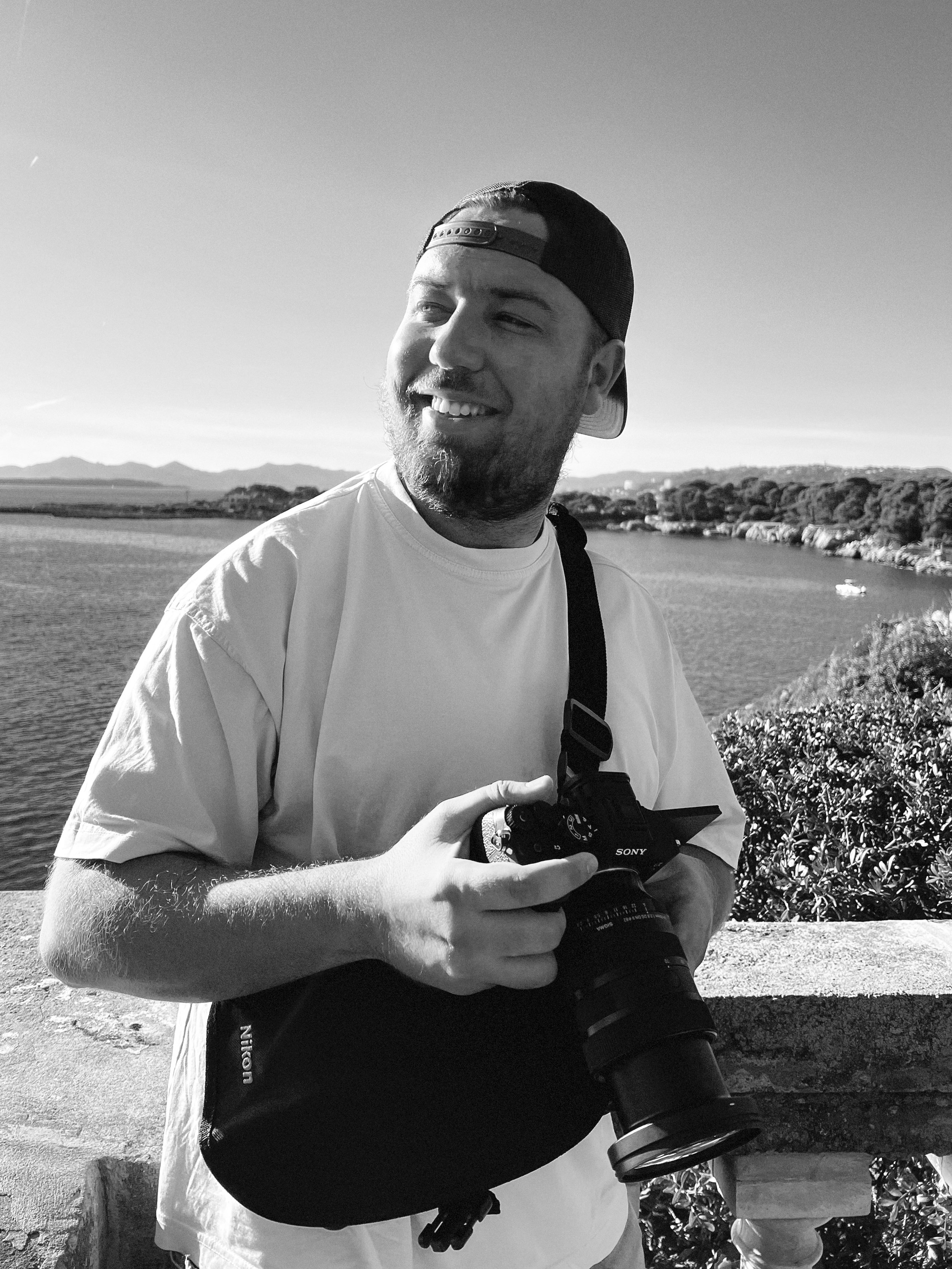Homme souriant portant un appareil photo, en extérieur avec un paysage d'eau et de montagnes en arrière-plan, en noir et blanc.