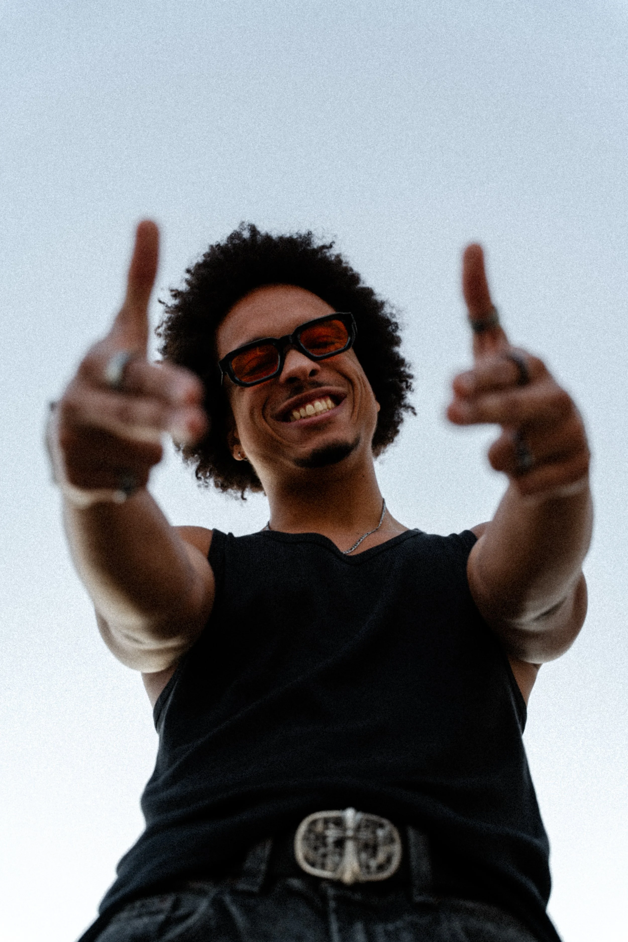 Jeune homme souriant portant des lunettes de soleil noires, un t-shirt noir, avec des bijoux, pointant vers la caméra, fond de ciel clair.