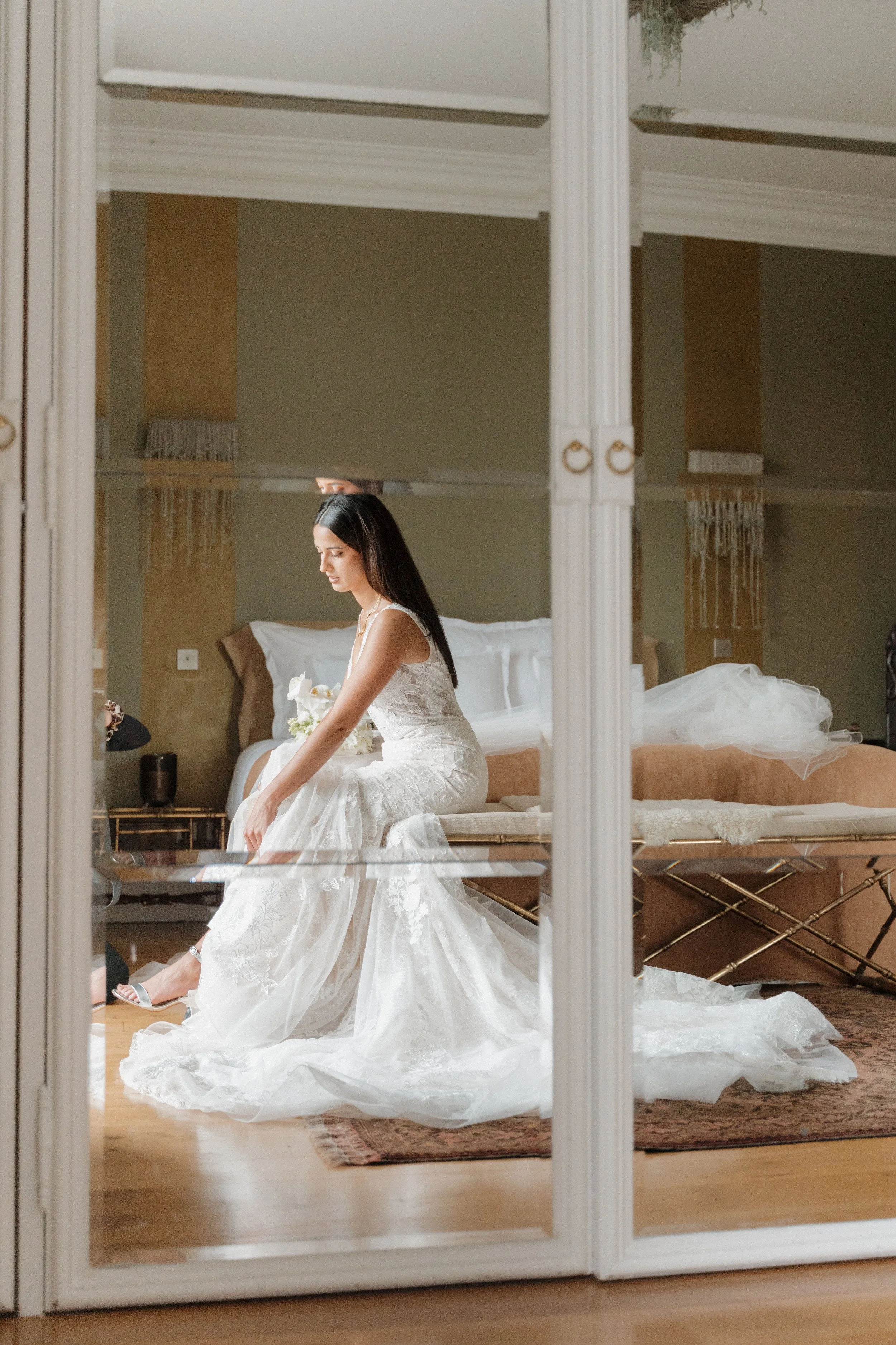 Une femme en robe de mariée blanche assise sur un lit, vue à travers une porte en miroir.  session femme mariées et robes haut de gamme, brindos, hôtel et lac,  photographe mariage bordeaux et Lyon et destination wedding
