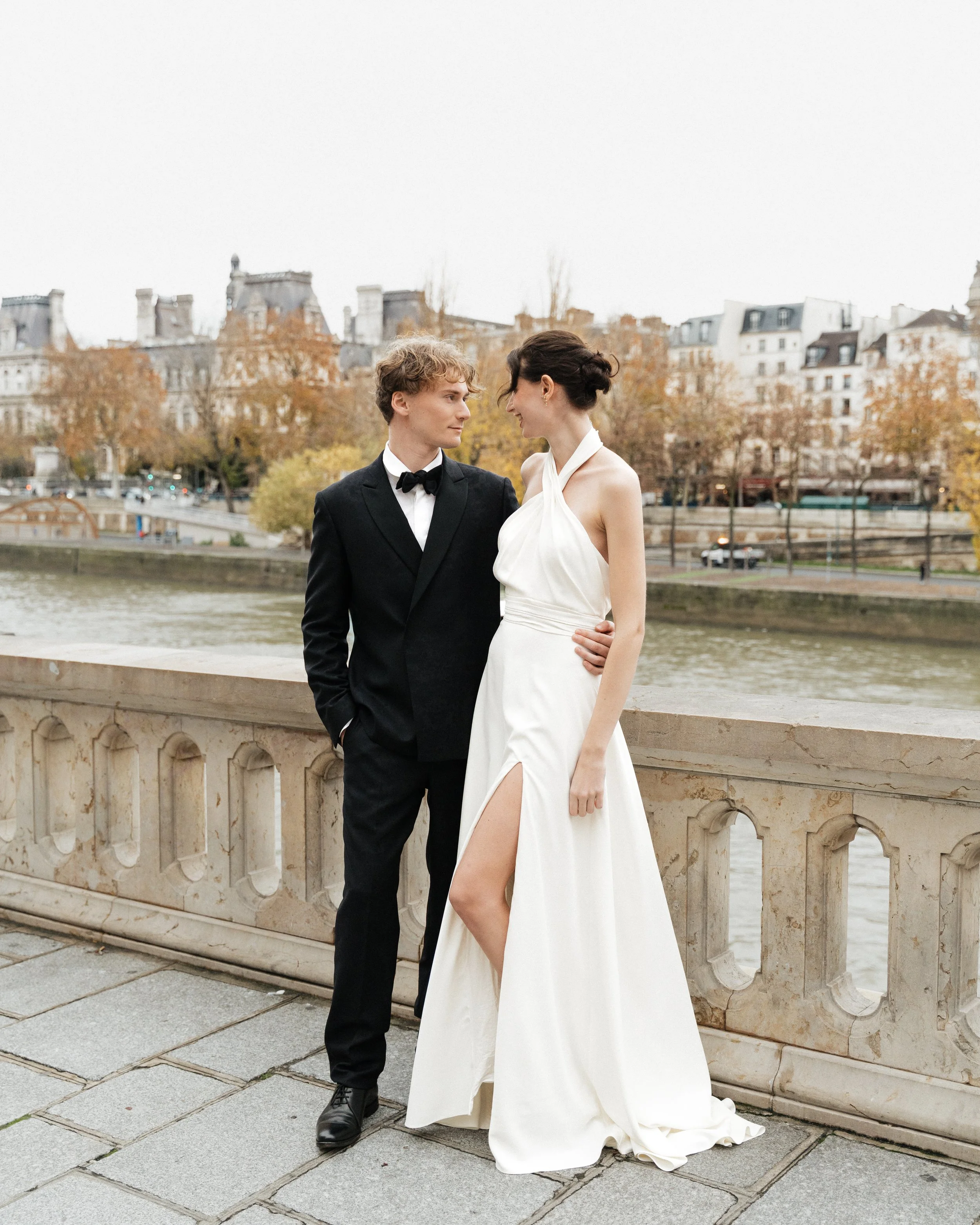 Un couple en tenue de mariage se tient sur un pont avec la rivière et des bâtiments parisiens en arrière-plan. Session engagement paris, Louvre, mariage haut de gamme, destination wedding. Robe de mariées, madame a dit oui