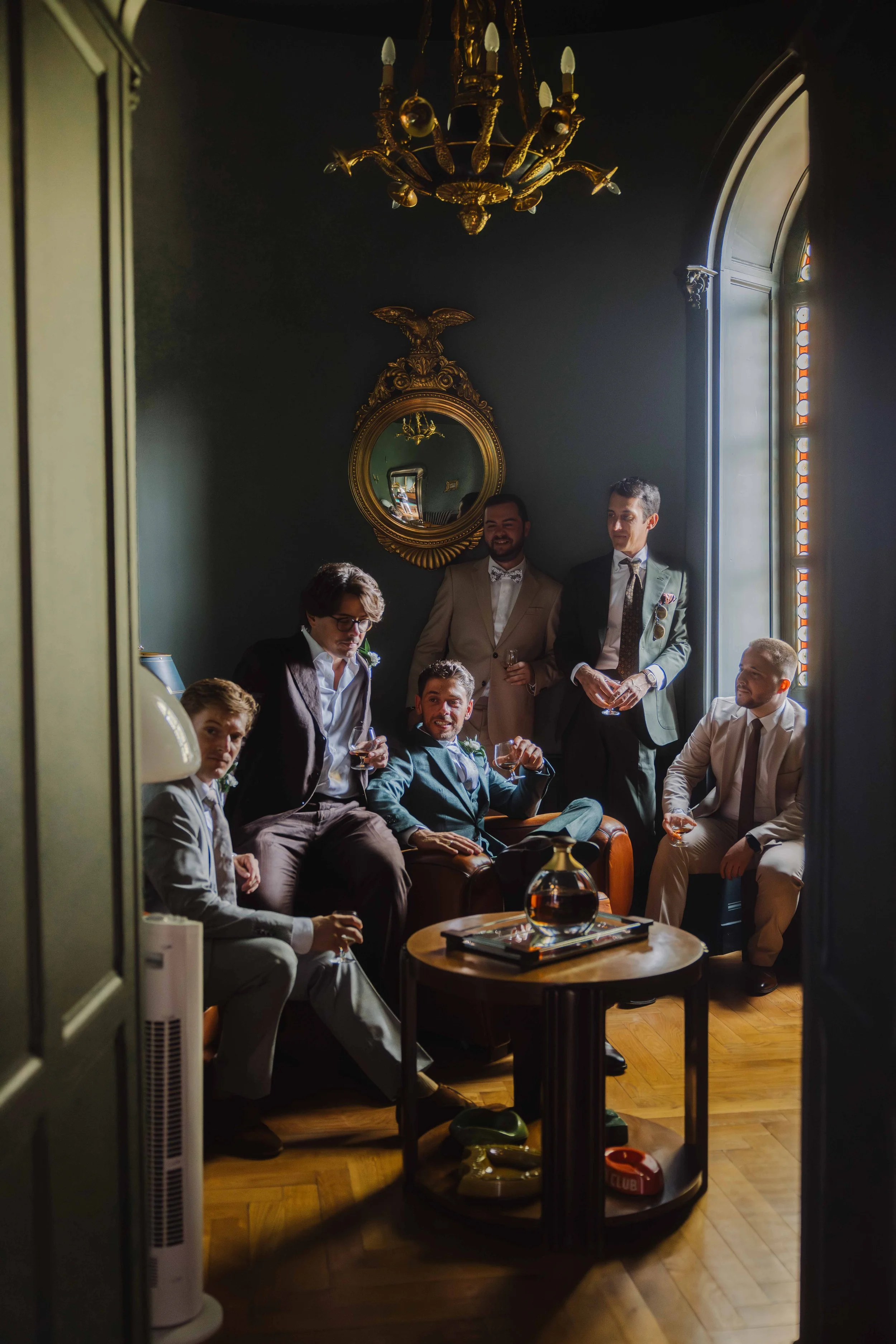 Groupe de sept hommes élégamment habillés, certains en costume clair et d'autres en costume foncé, posant dans une pièce sombre avec une lumière naturelle venant d'une fenêtre grande et arquée, décorée de moulures et d'un grand miroir doré. 