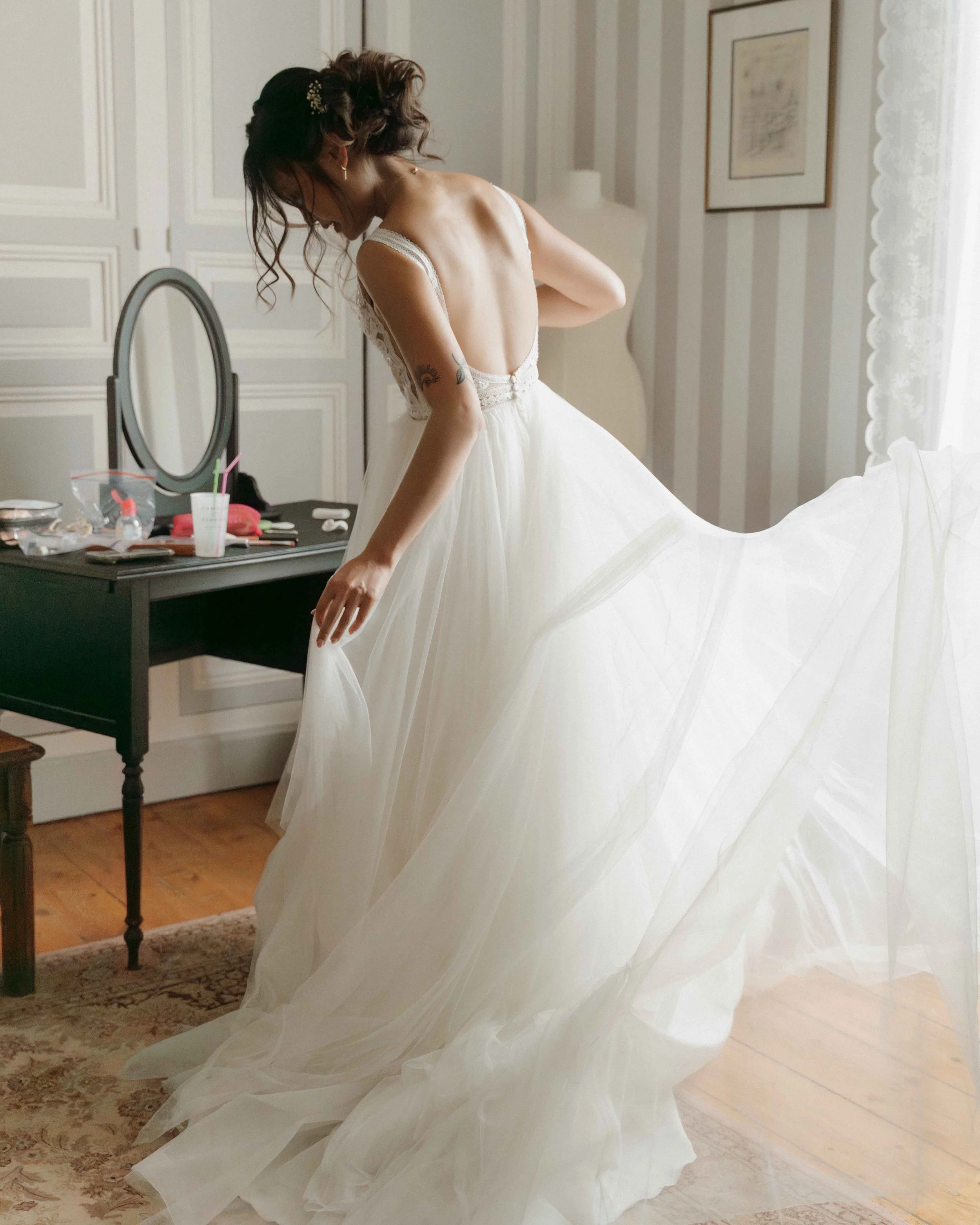 Femme en robe de mariée blanche dans une pièce lumineuse, ajustant la robe devant un miroir. Domaine de tourieux à Savigny dans le beaujolais à Lyon. Mariage cérémonie laïque, photographe Lyon haut de gamme, duo photographe, argentique.