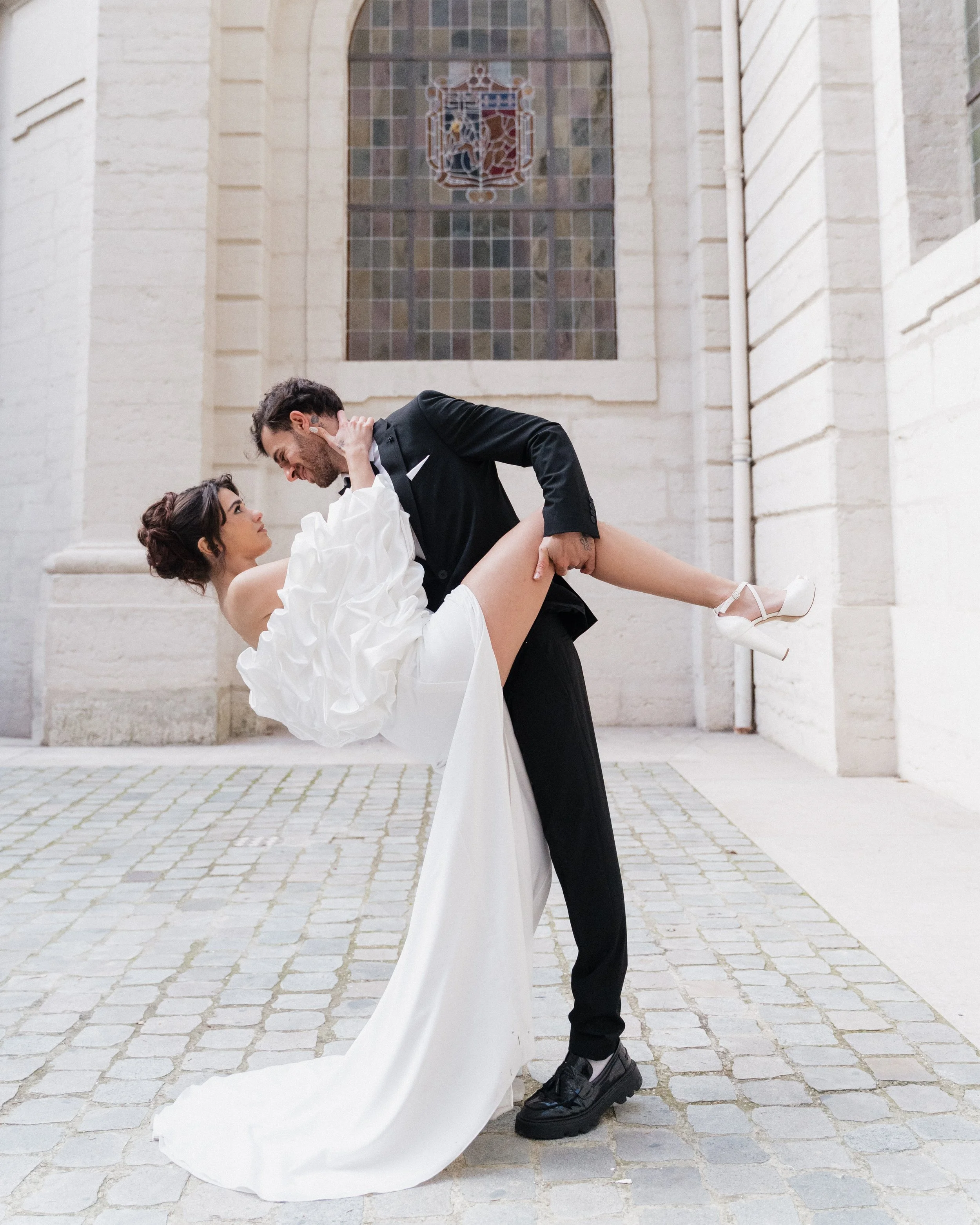 Un couple en robe de mariage et costume danse devant un bâtiment en pierre avec une grande fenêtre en verre coloré à l'hôtel dieu à Lyon 2ème, à l'intercontinental, session engament.