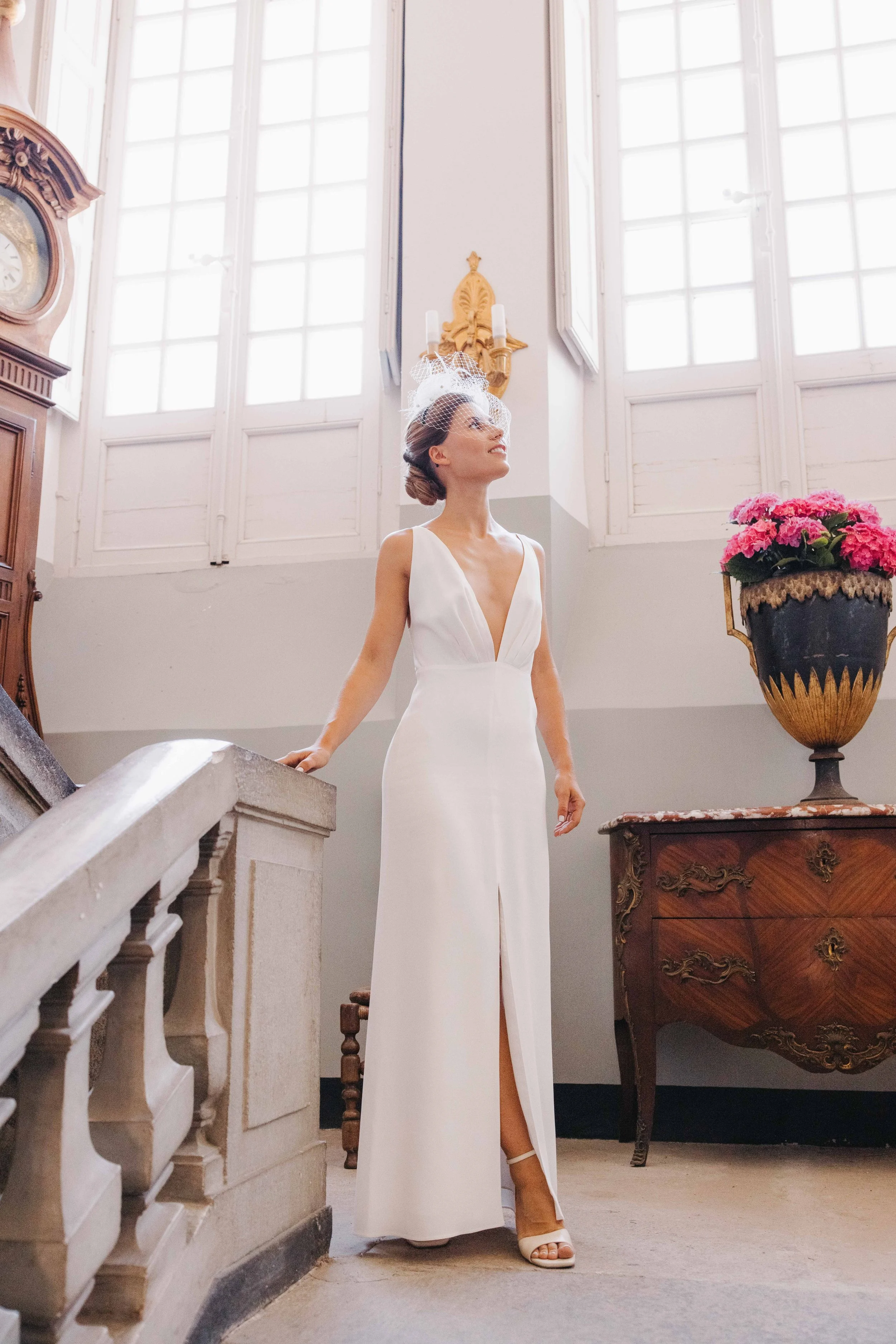 Femme en robe de mariée blanche élégante avec un décolleté profond et une fente, portant un voile avec un filet en tulle et des chaussures à talons, debout dans une pièce lumineuse avec de hautes fenêtres, une horloge ancienne et un grand vase de fle