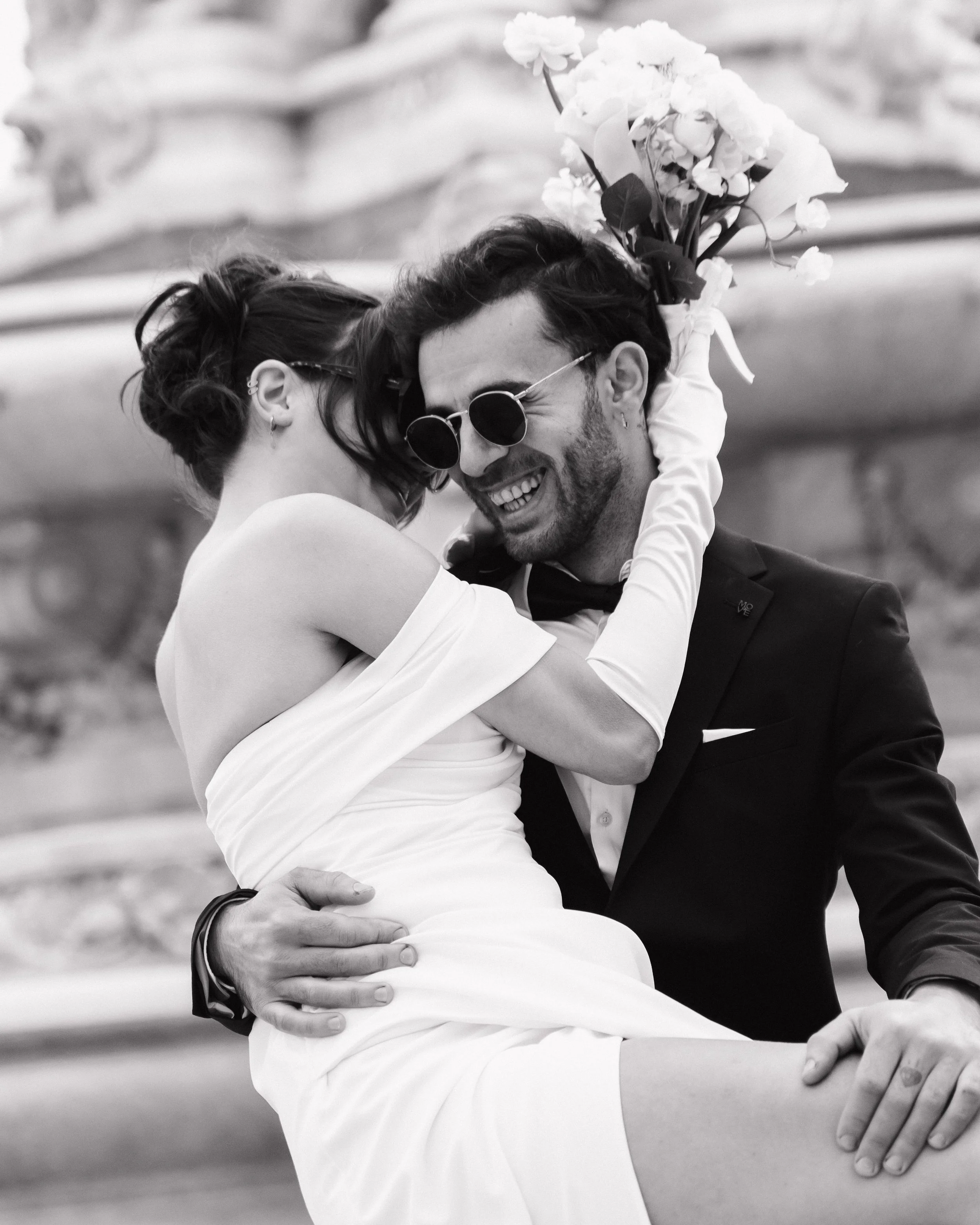 Un couple de mariés, l'homme portant un smoking et la femme une robe blanche, partage un moment heureux avec la femme tenant un bouquet de fleurs, tous deux souriants et en train de s'étreindre. à Lyon place des jacobins, photographe duo 
