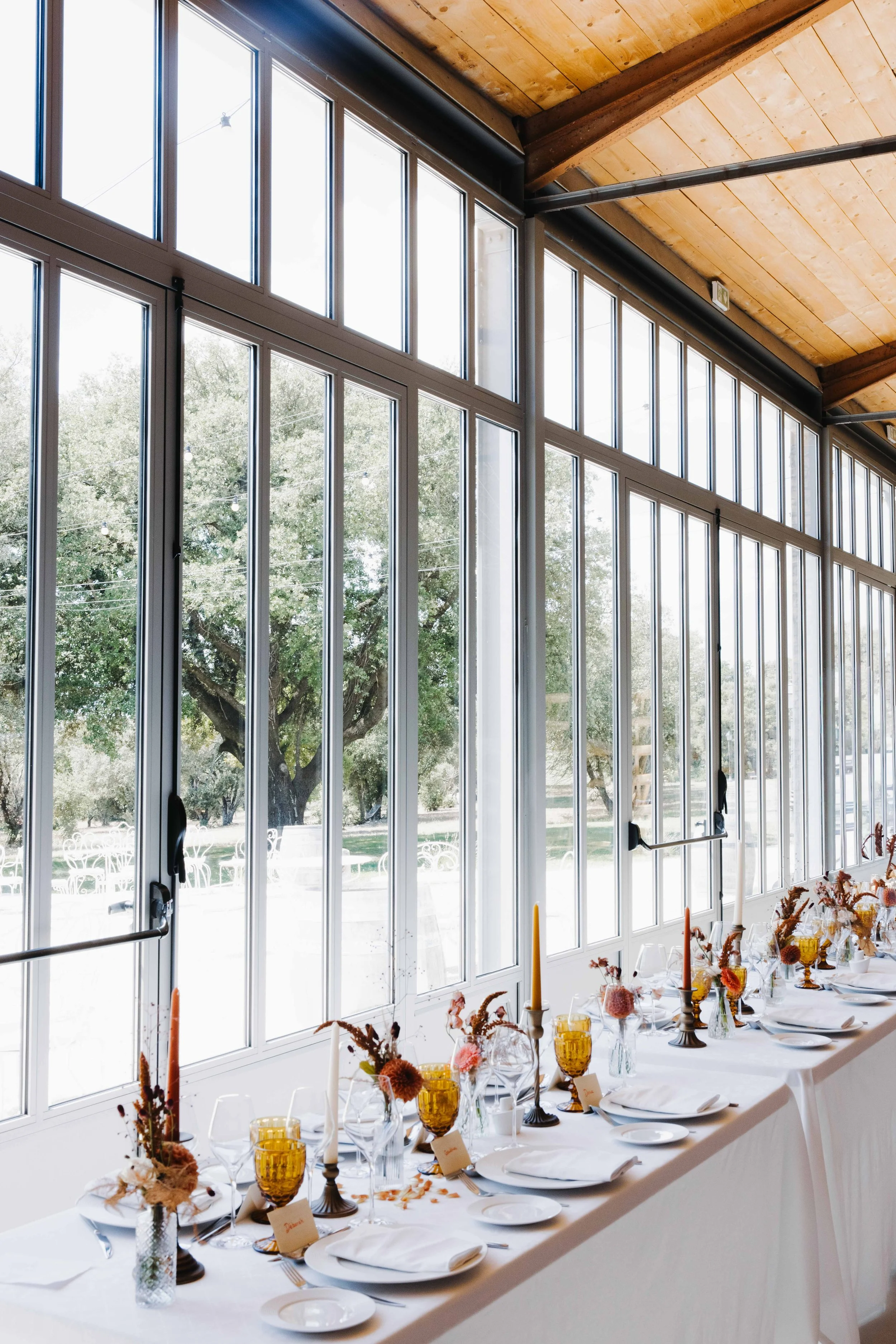 Une grande table décorée avec des fleurs, bougies et vaisselle, située devant de grandes fenêtres laissant passer beaucoup de lumière naturelle dans une pièce avec plafond en bois. Domaine du bijoutier à grignan, drome provençal, traiteur mariage 