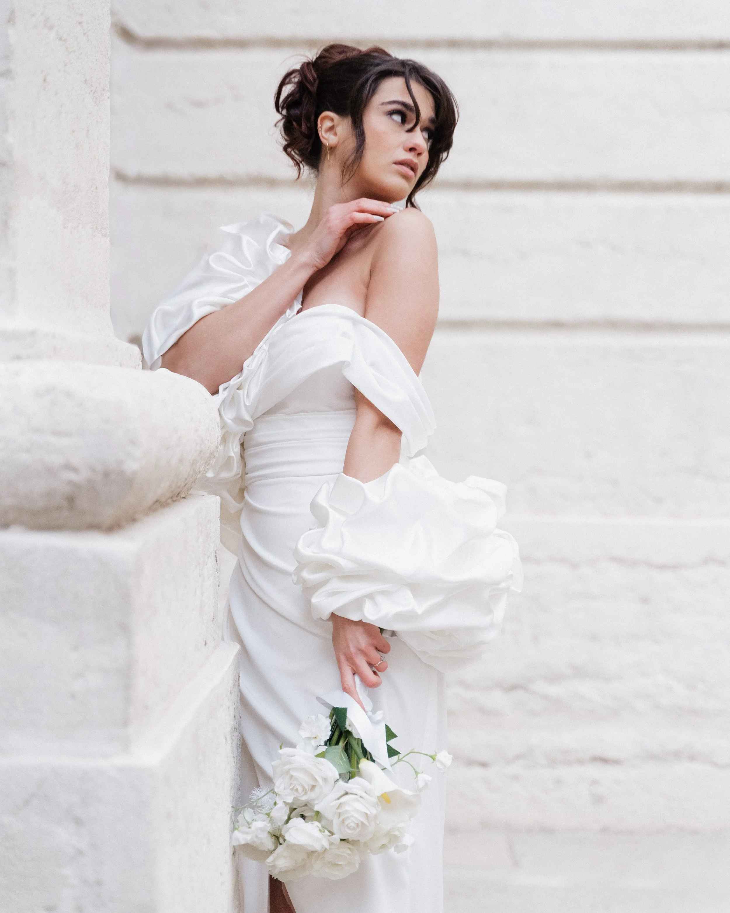 Une femme vêtue d'une robe blanche tient un bouquet de fleurs blanches, se tenant près d'un mur en pierre, avec une expression pensive à l'hôtel dieu à Lyon 2ème, à l'intercontinental, session engament.