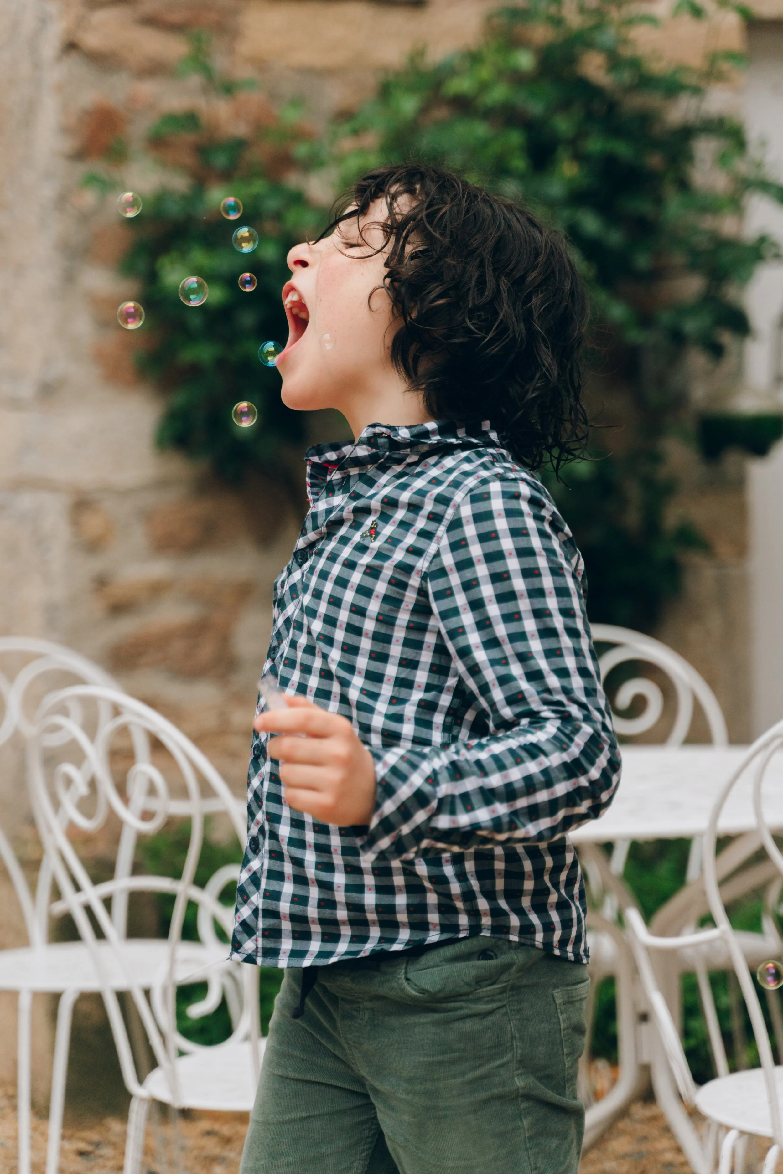 Un enfant avec des cheveux bouclés courts, portant une chemise à carreaux, joue avec des bulles de savon à l'extérieur, avec des chaises en fer blanc et un mur en pierre en arrière-plan. Domaine de tourieux à Savigny dans le beaujolais à Lyon. 