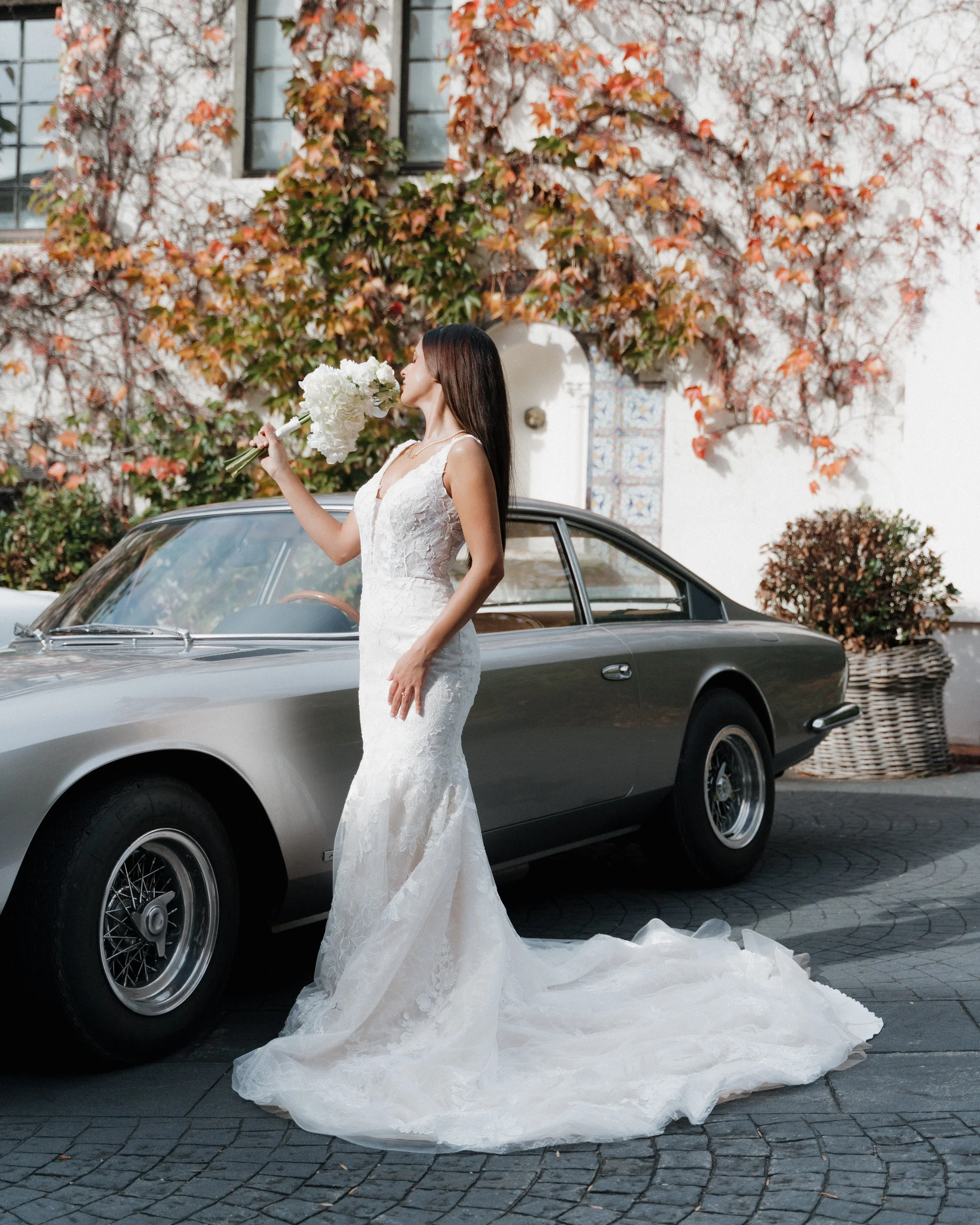 Une femme en robe de mariée blanche tenant un bouquet de fleurs blanches près d'une voiture vintage argentée, avec un mur couvert de feuillage aux couleurs d'automne en arrière-plan.  session femme mariées et robes haut de gamme, brindos hôtel 