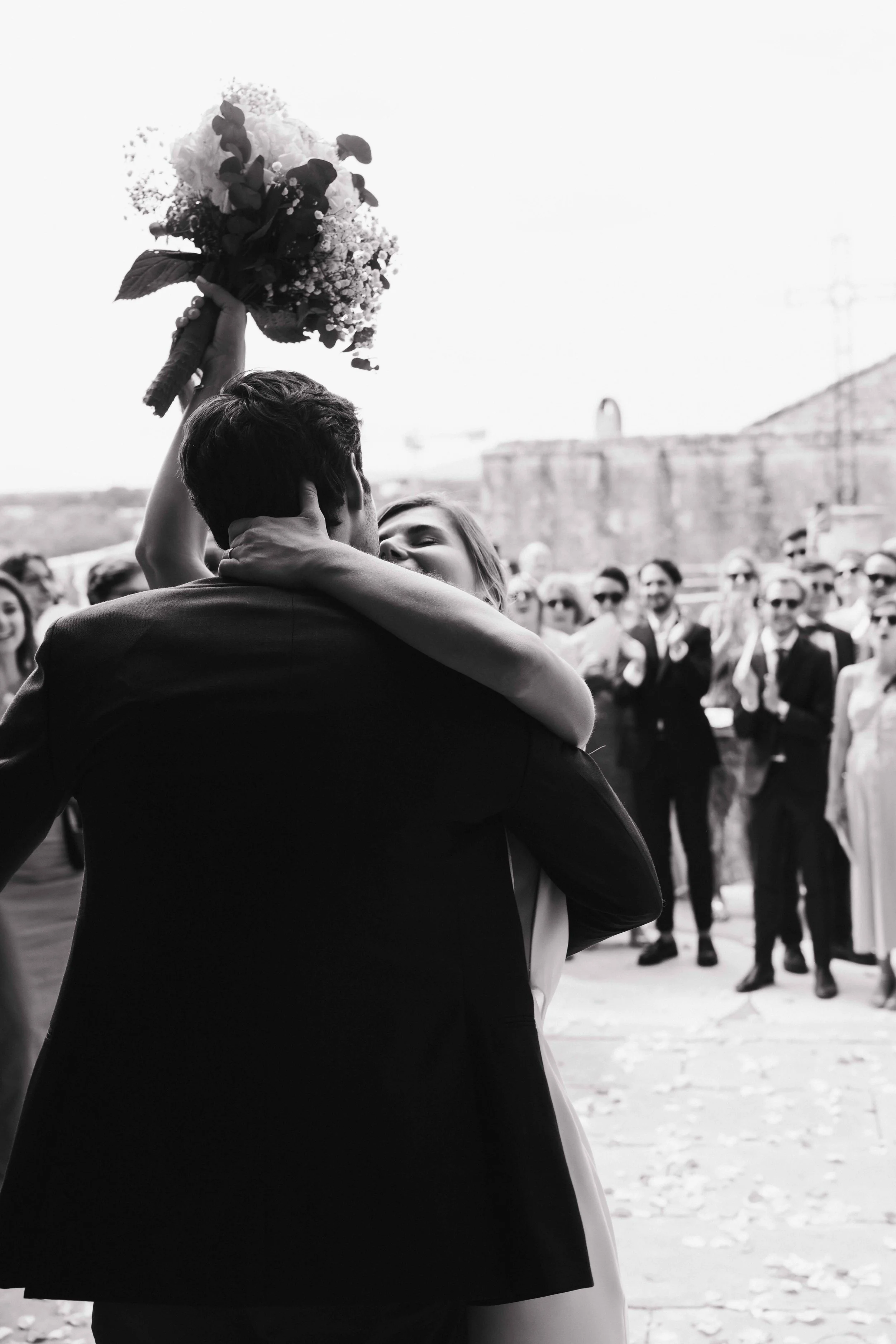 Un couple s'embrasse lors d'une réception de mariage en plein air, avec le marié tenant un bouquet de fleurs levé en l'air, et des invités en arrière-plan regardant la scène. Domaine du bijoutier à grignan, drome provençal, cérémonie religieuse 