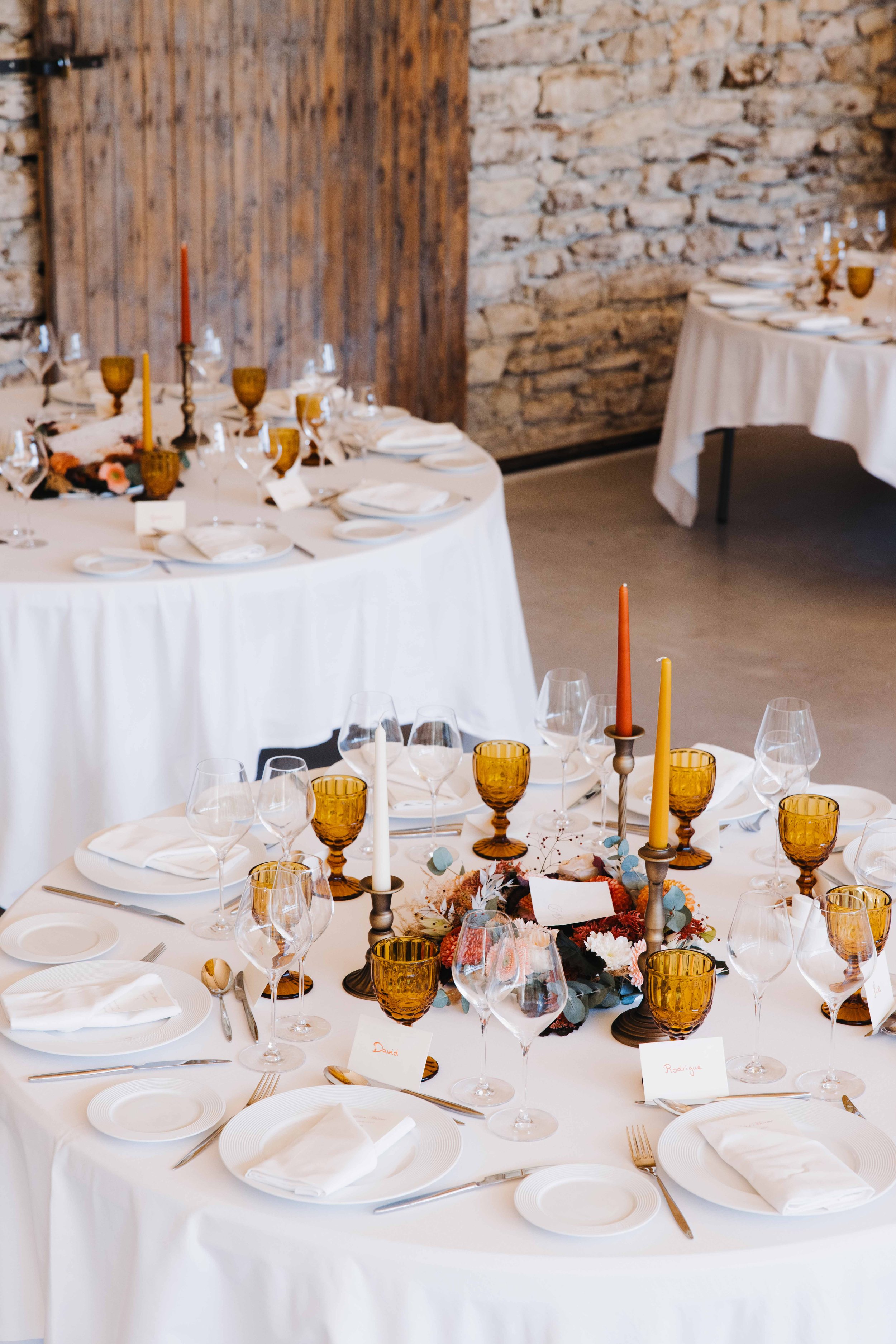 Table de réception élégante avec vaisselle blanche, verres en verre et en verre ambré, bougies colorées, fleurs, dans une ambiance rustique avec des murs en pierre et un bois visible. Domaine du bijoutier à grignan, drome provençal, traiteur mariage 