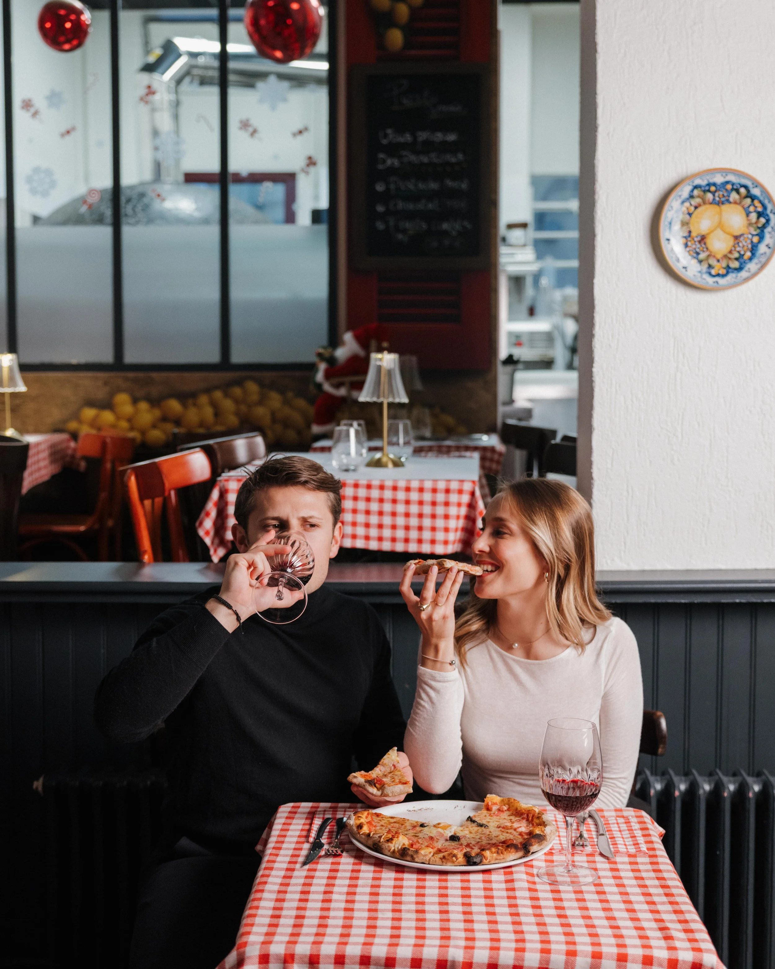 Un jeune homme et une jeune femme assis à une table de restaurant, partageant une pizza, la femme tenant une part de pizza et le jeune homme buvant du vin rouge, avec un décor de restaurant italien à l'arrière-plan. session engagement couple Lyon 