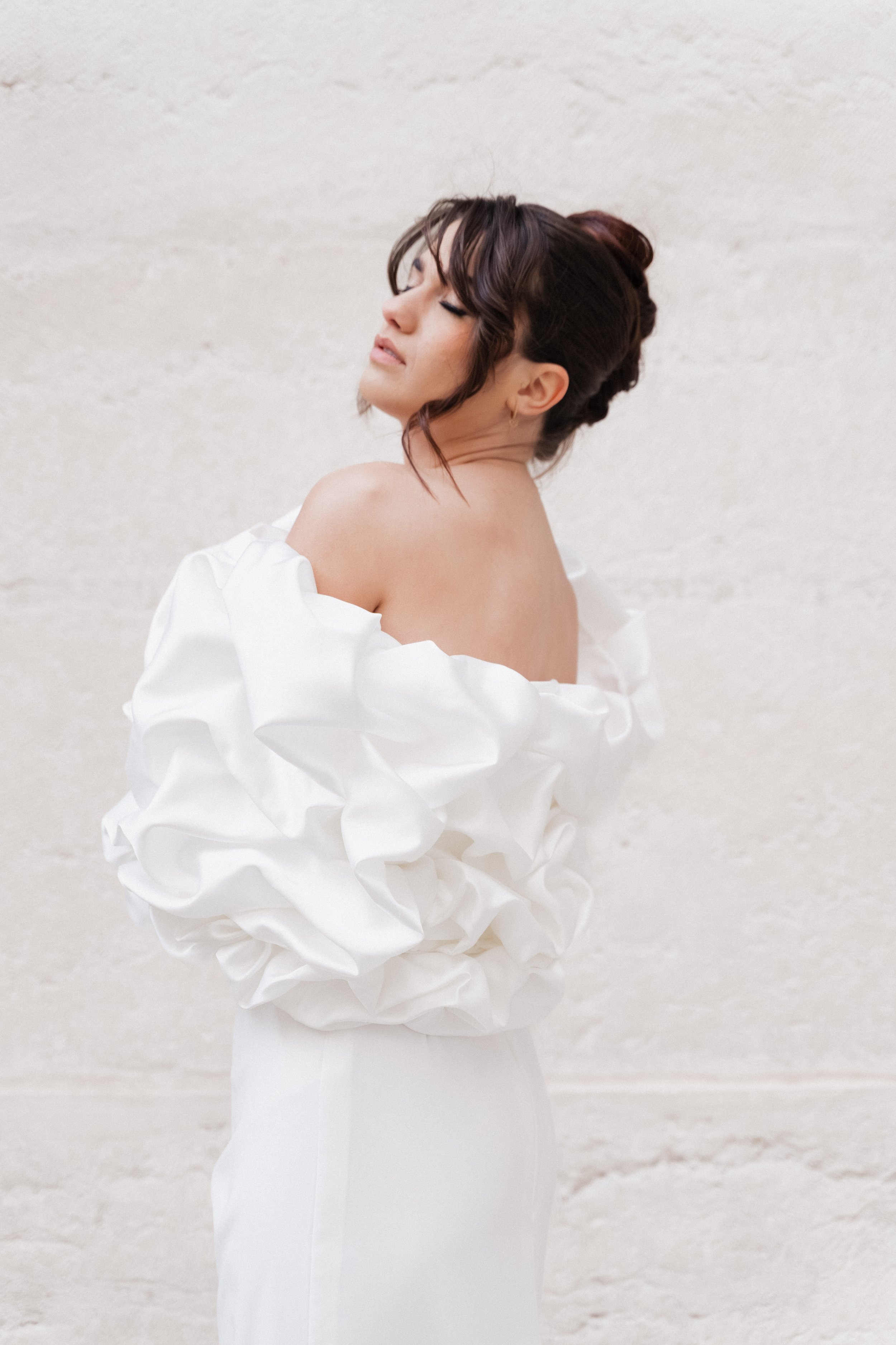 Femme portant une robe blanche avec des manches bouffantes et une robe sans épaules, posant avec les yeux fermés contre un mur beige  à l'hôtel dieu à Lyon 2ème, à l'intercontinental, session engament, photographe mariage duo haut de gamme.