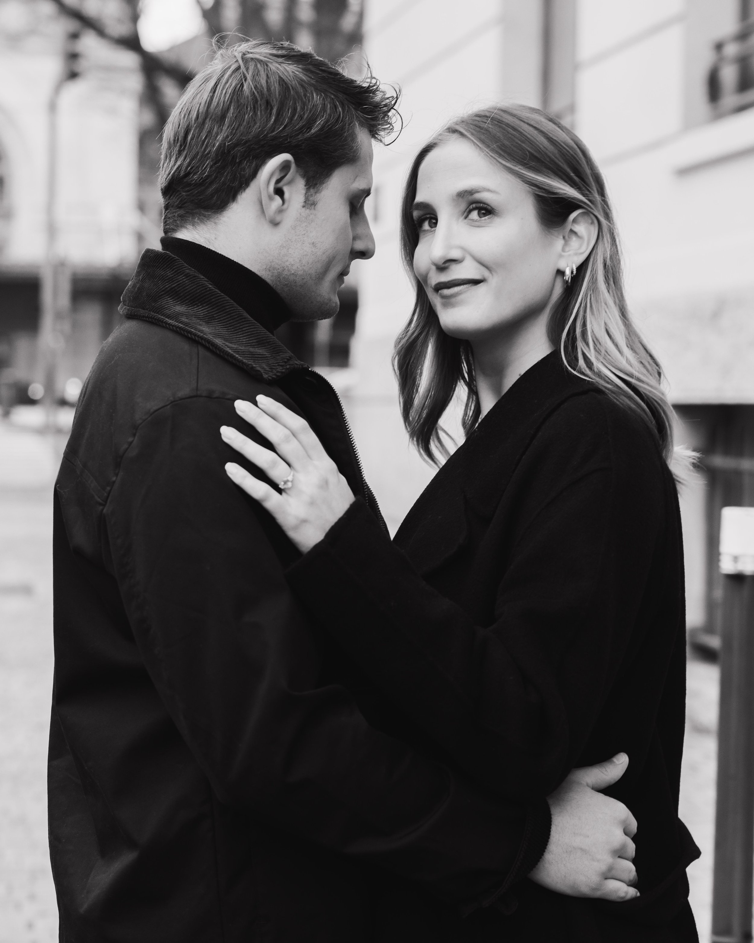 Un couple s'embrasse et se regarde dans une rue urbaine, l'homme porte une veste sombre, la femme a des cheveux longs et porte une veste noire. session engagement couple Lyon 6eme, brotteaux, photographe mariage bordeaux et Lyon et destination