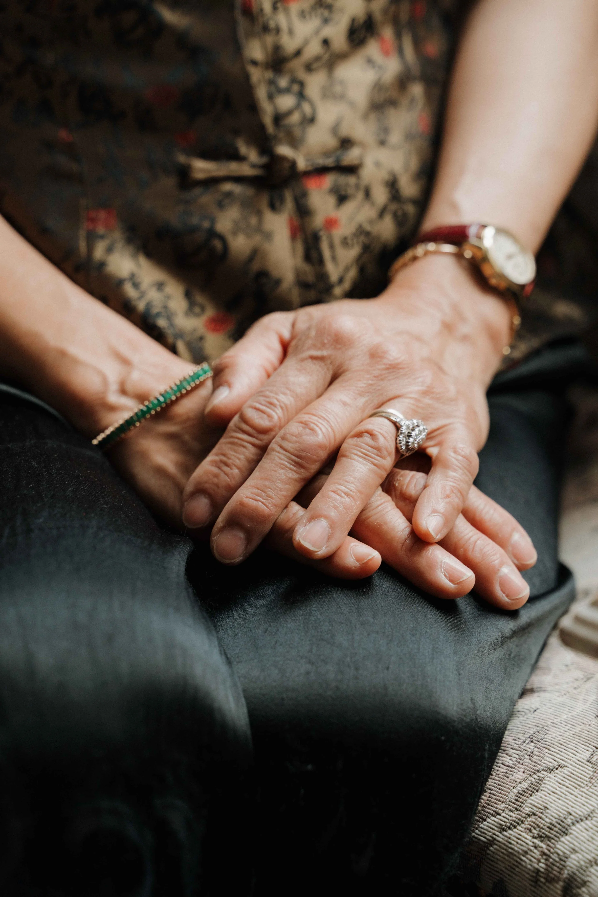 Mains croisées d'un couple, avec une bague de mariage, portant des bracelets, reposant sur la jambe d'une personne portant un vêtement à motif floral. Domaine de tourieux à Savigny dans le beaujolais à Lyon. Mariage cérémonie laïque, photographe Lyon