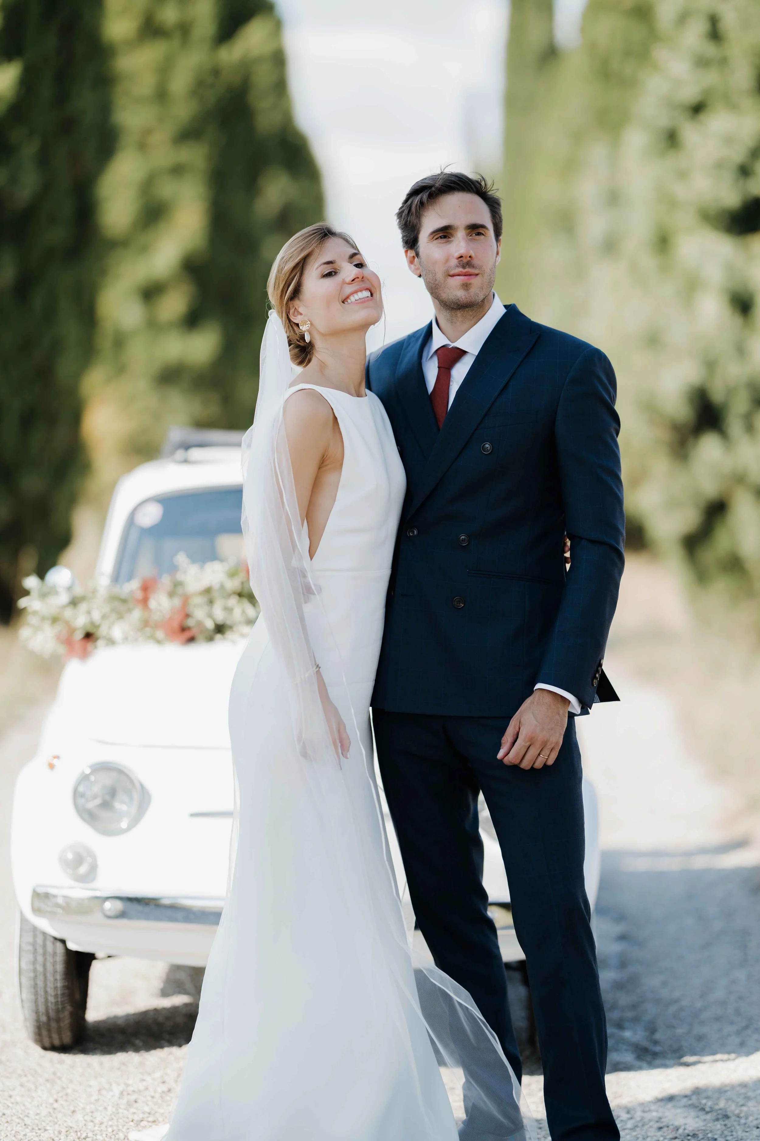 Un couple de mariés en costume et robe de mariée, posant ensemble devant une voiture décorée de fleurs, dans un environnement nature avec des arbres. Domaine du bijoutier à grignan, drome provençal, coucher de soleil, session couple, fiat 500