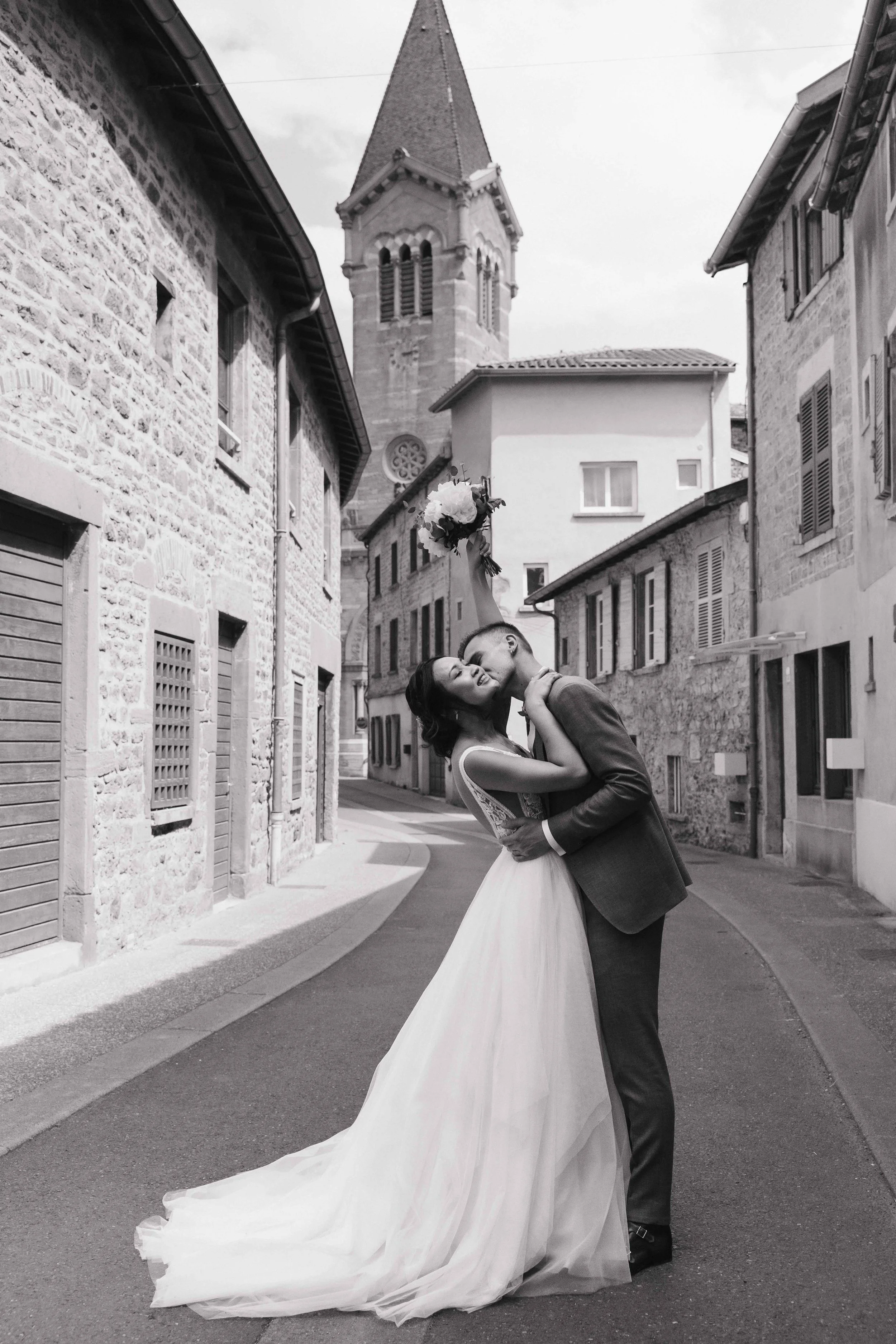 un couple de mariés embrassant dans une rue pavée avec des bâtiments en pierre et une église en arrière-plan. Domaine de tourieux à Savigny dans le beaujolais à Lyon. Mariage cérémonie laïque, photographe Lyon haut de gamme, duo photographe, film