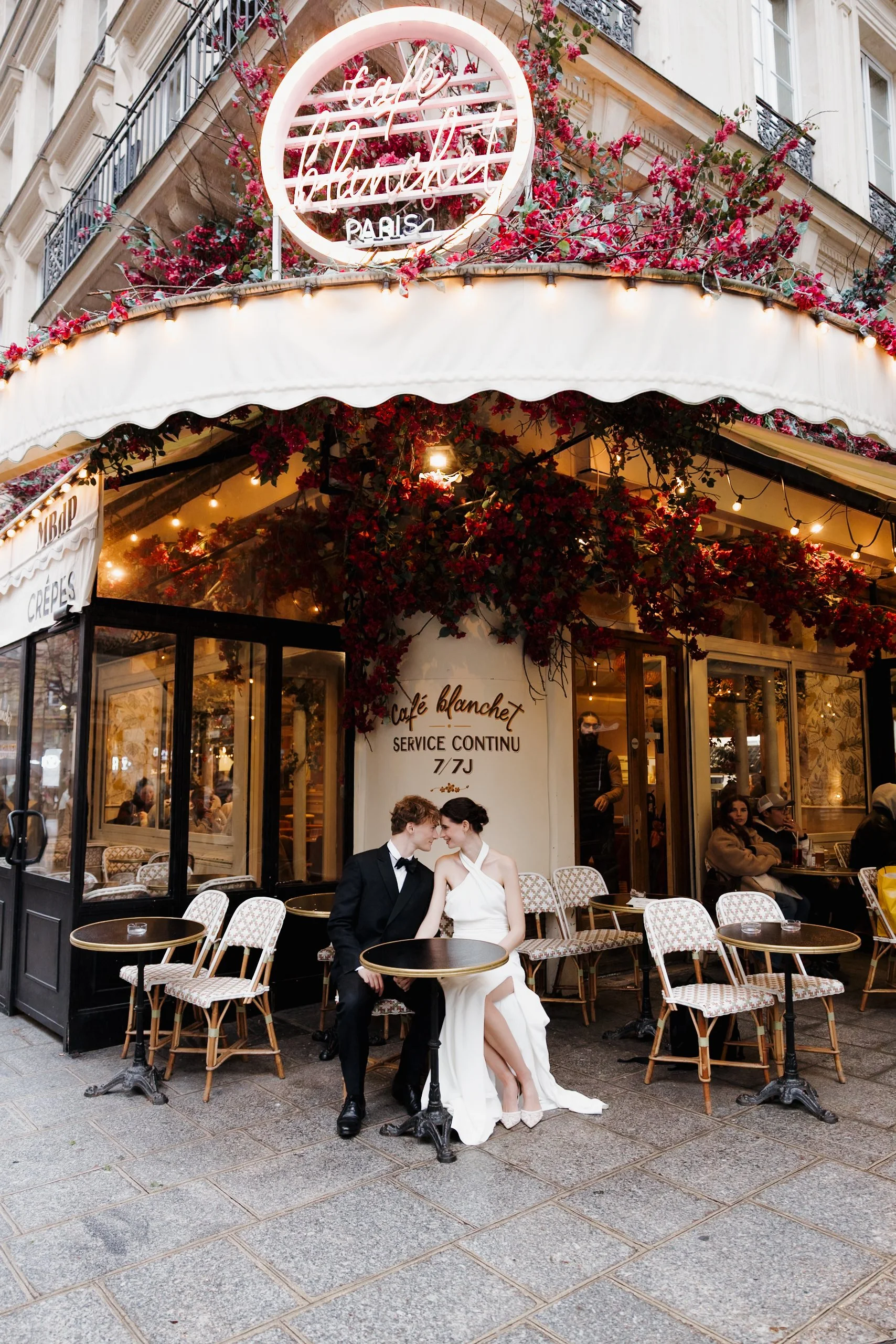 Un couple habillé en mariage, assis à une table devant un café parisien, dans une ambiance chaleureuse et romantique, avec des décorations florales et des lumières. Session engagement café parisien, photographe mariage paris haut de gamme 
