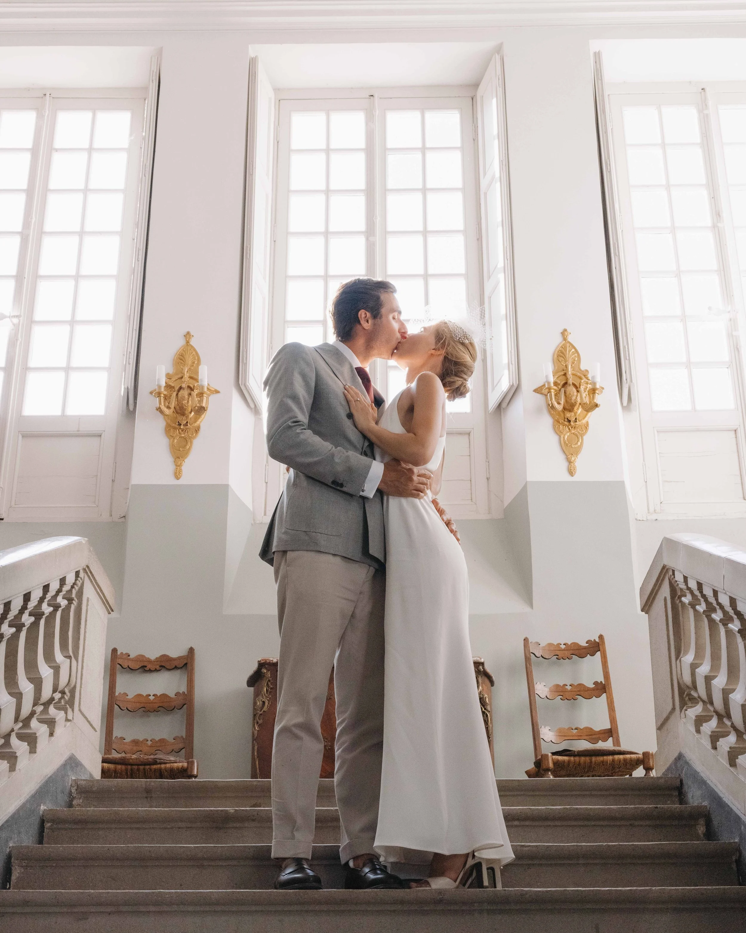 Un couple en mariage s'embrassent au sommet d'un escalier dans une grande pièce lumineuse avec de grandes fenêtres et des murs blancs. Mariage au chateau de lachal dans le rhone alpes à côté de Lyon. Cérémonie civil et laïque. Robe de mariées 