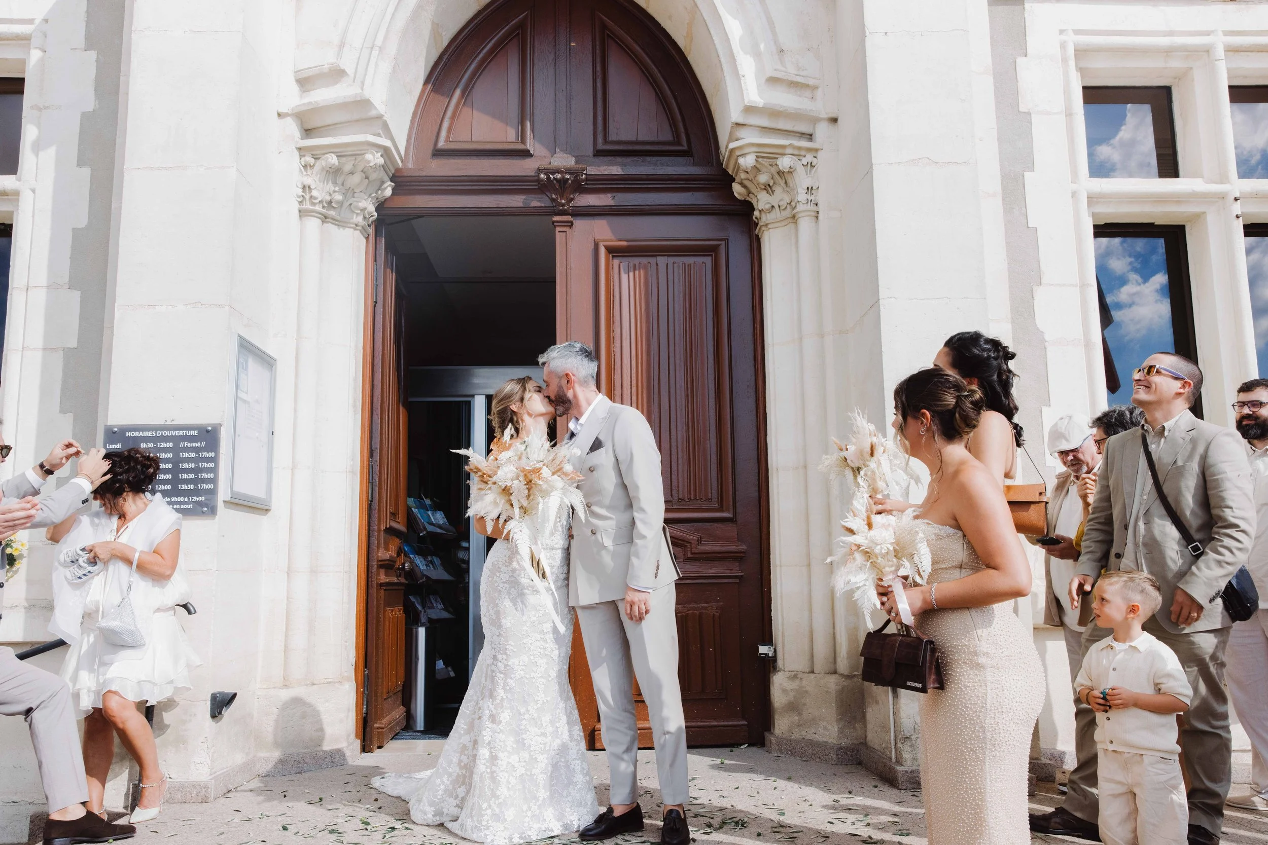 Un couple en mariage s'embrasse devant une église, entouré de leurs invités. Les femmes portent des robes élégantes, les hommes des costumes. Il fait beau, et la scène est festive. Photographe mariage Isère, ferme du rocher, cérémonie civil et laïque