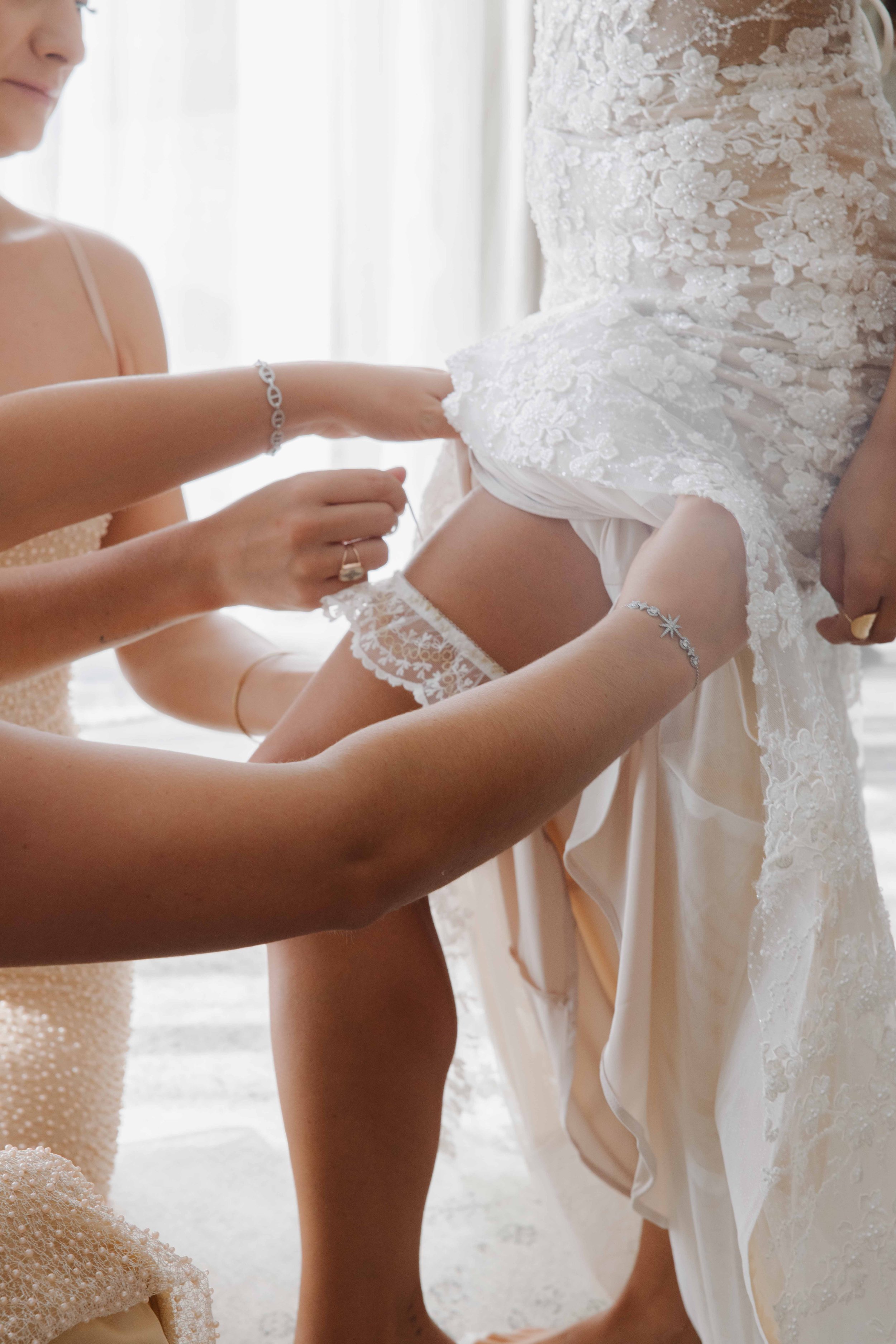 Une femme en robe de mariée blanche est aidée à ajuster sa robe par deux autres femmes. La scène se passe en intérieur, près d'une fenêtre, avec un éclairage doux. Photographe mariage Isère, ferme du rocher, cérémonie civil et laïque. Lyon 
