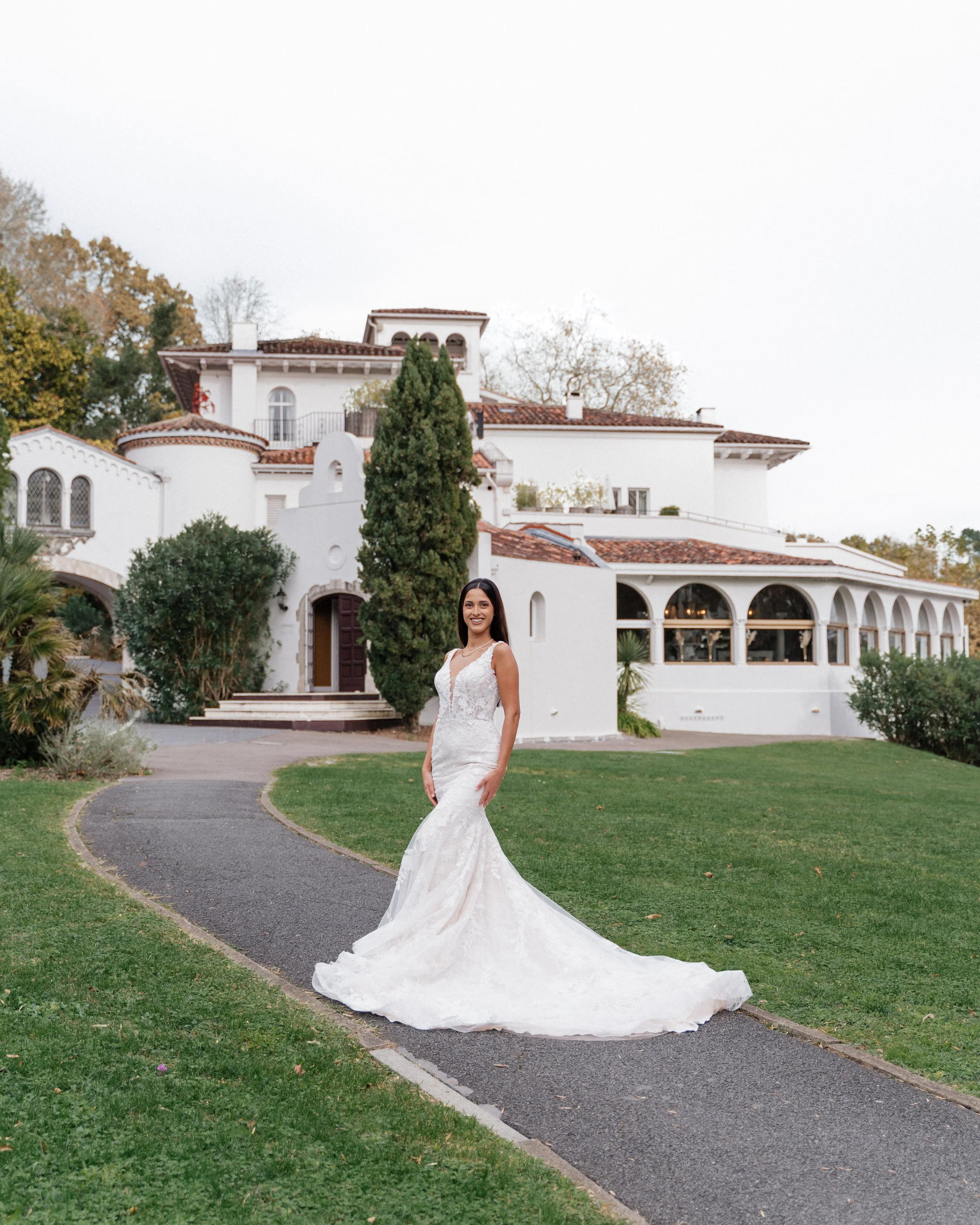Une femme en robe de mariée blanche se tient sur un chemin menant à une grande maison blanche dans un jardin verdoyant.  session femme mariées et robes haut de gamme, brindos, hôtel et lac,  photographe mariage bordeaux et Lyon et destination wedding