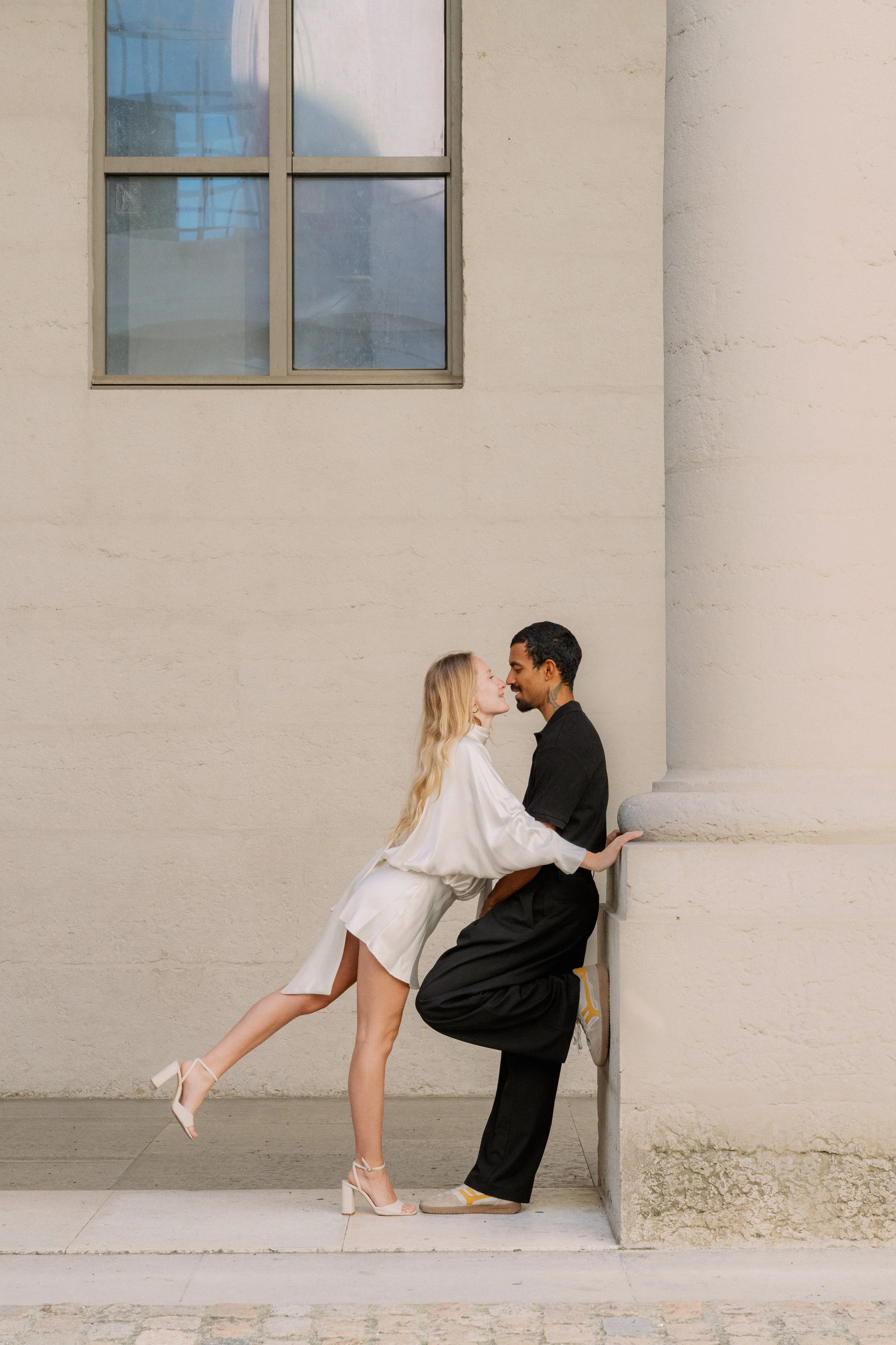 Un couple debout contre un mur beige, la femme en robe blanche et chaussures à talons roses, le homme en pantalon noir, t-shirt noir et chaussures beige, posant près d'une colonne, avec une fenêtre en haut à gauche à l'hôtel dieu à Lyon 2ème