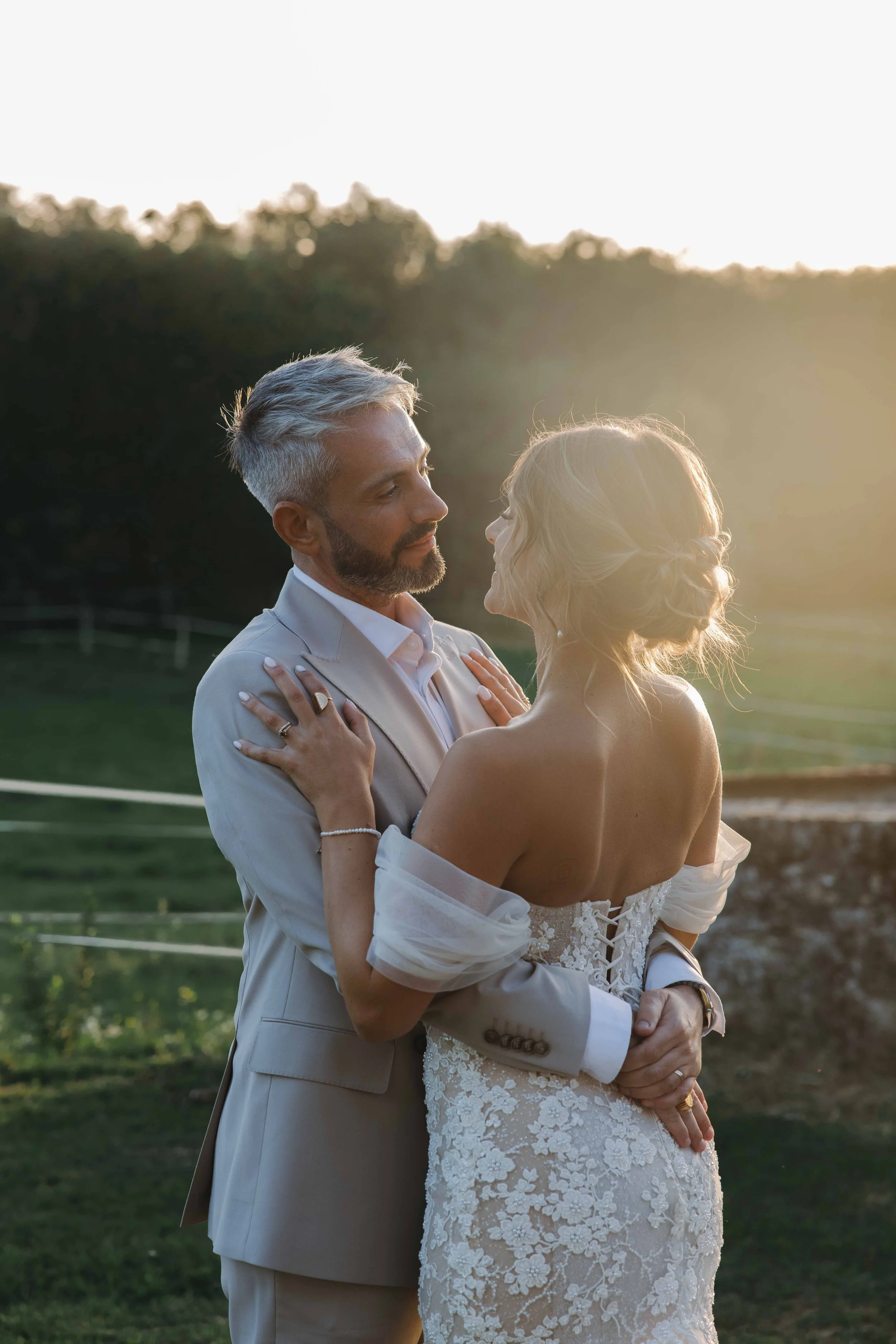 Un couple en tenue de mariage se tenant dans un champ avec une forêt en arrière-plan, au coucher du soleil. Photographe mariage Isère, ferme du rocher, cérémonie civil et laïque. Bordeaux et Lyon et destination wedding. Photos de couple, session coup