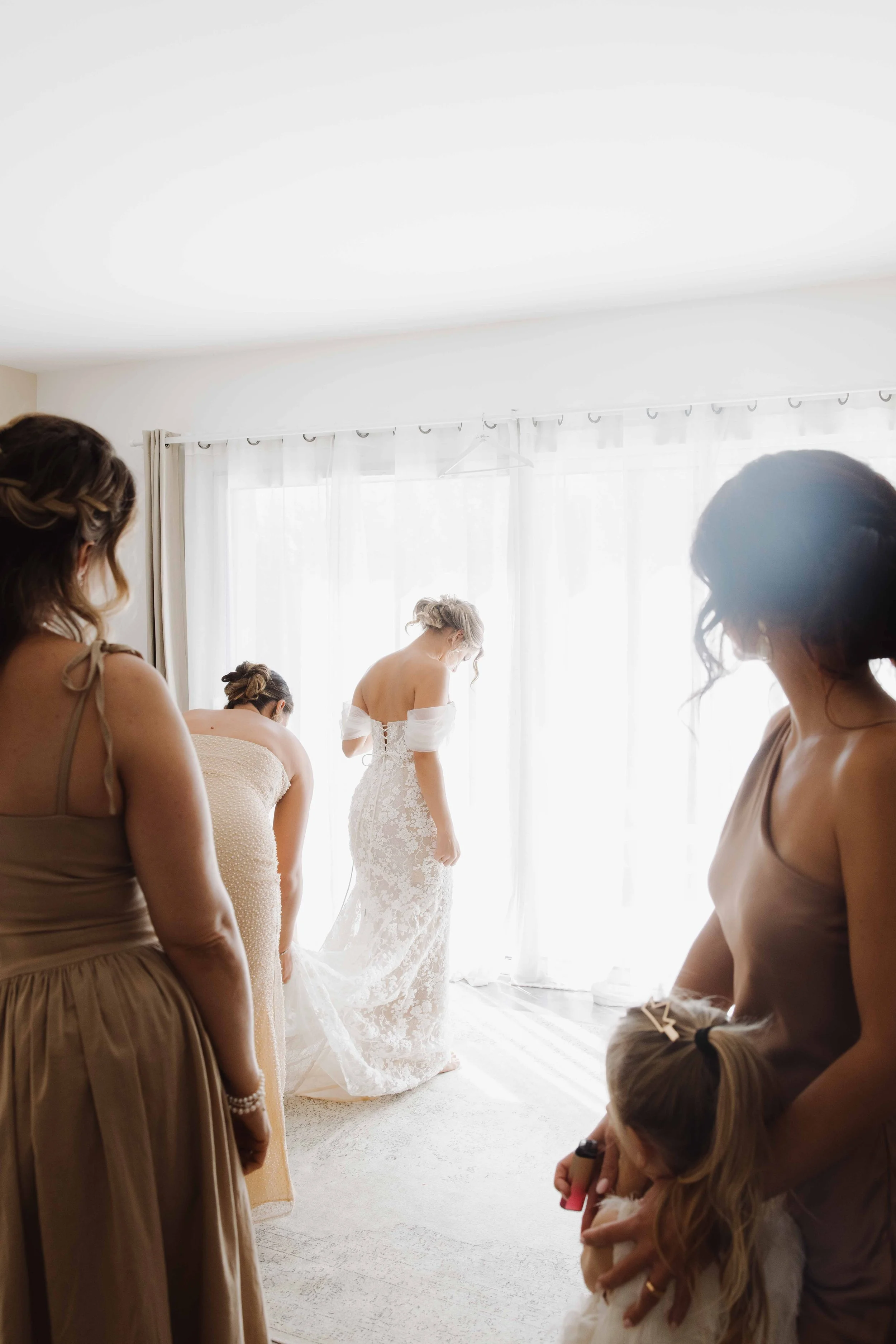 Une mariée en robe de mariée blanche, entourée de plusieurs femmes habillées en beige ou taupe, se prépare dans une pièce lumineuse avec des rideaux translucides en arrière-plan. Photographe mariage Isère, ferme du rocher, cérémonie civil et laïque. 