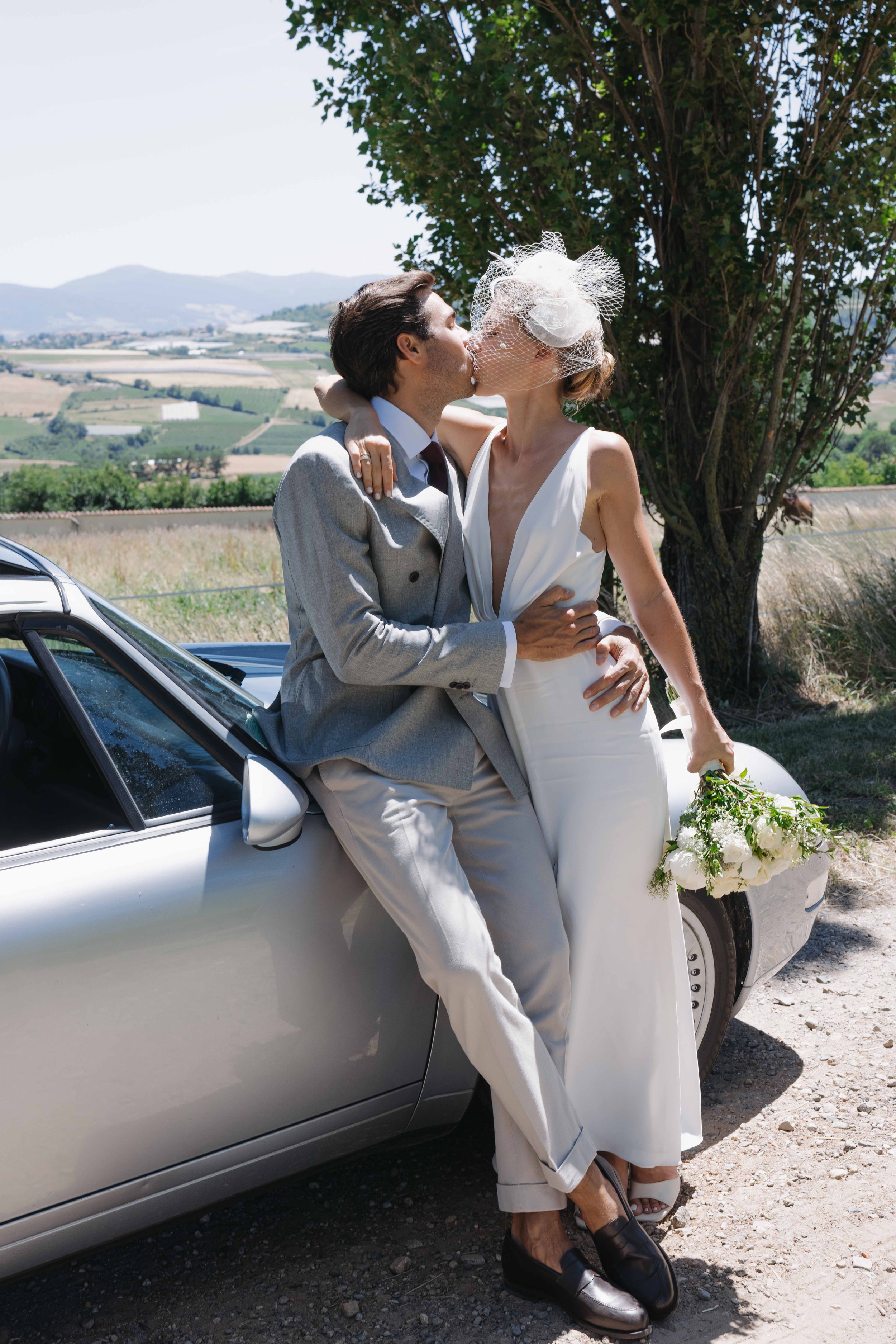 Un couple en vêtements de mariage s'embrassent devant une voiture avec un paysage rural en arrière-plan, la femme tient un bouquet de fleurs blanches. Mariage au chateau de lachal dans le rhone alpes à côté de Lyon. Cérémonie civil et laïque.