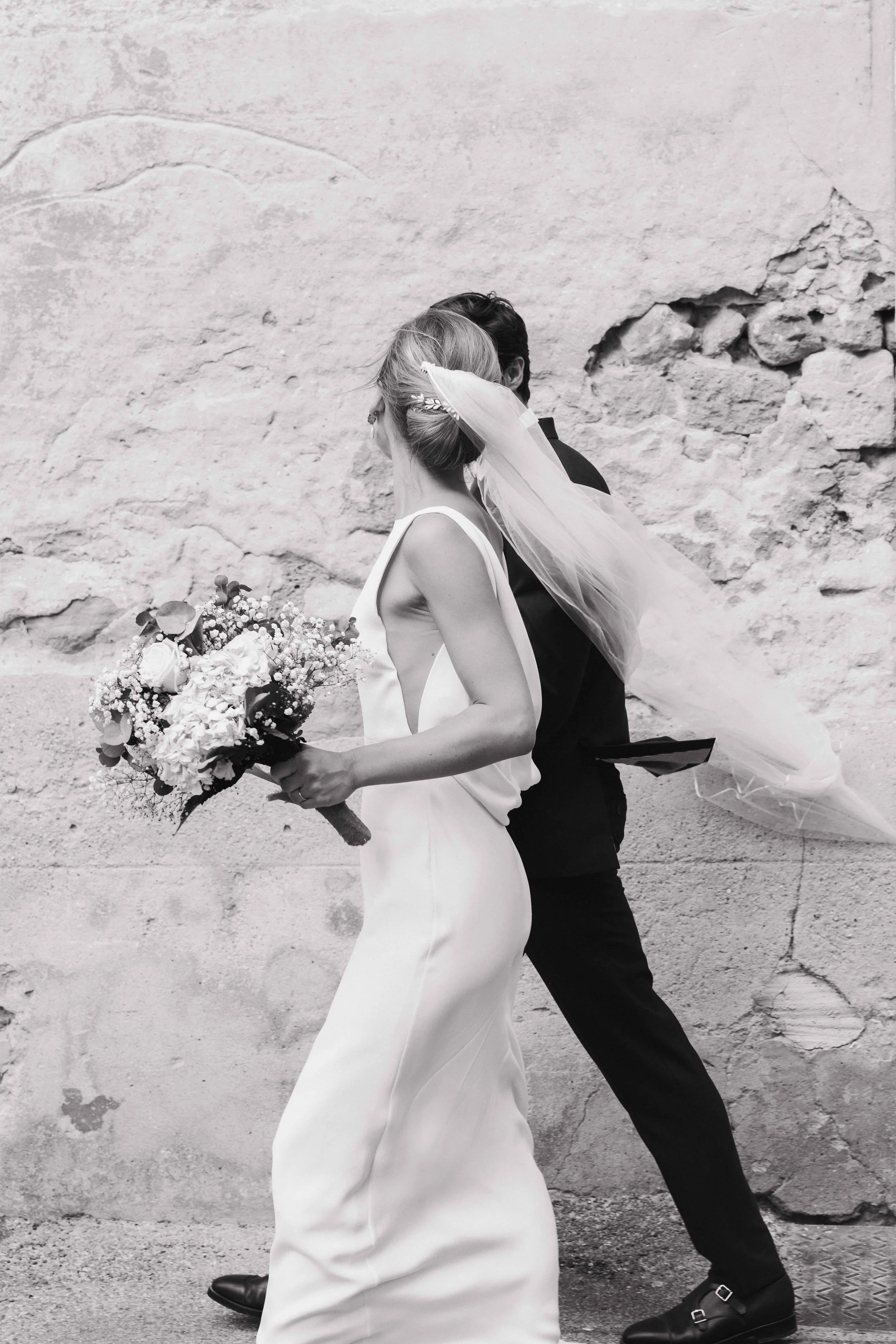 Un couple de mariés marche côte à côte contre un mur en pierre, la femme porte une robe blanche et tient un bouquet de fleurs, la tête couverte d'un voile, l'homme porte un costume noir. Domaine du bijoutier à grignan, drome provençal, église grignan