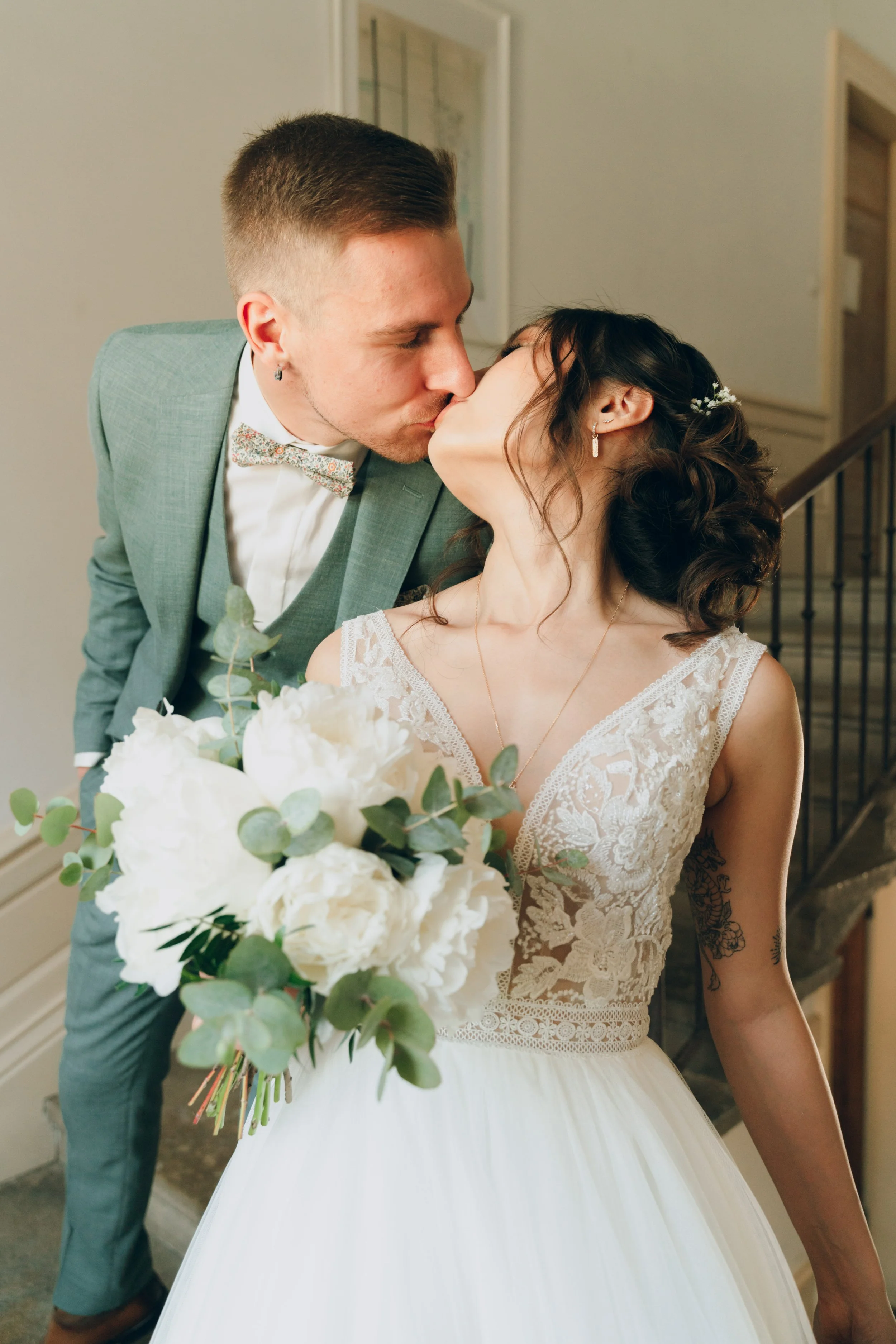 Un couple en tenue de mariage partage un baiser, la femme tient un bouquet de fleurs blanches. Domaine de tourieux à Savigny dans le beaujolais à Lyon. Mariage cérémonie laïque, photographe Lyon haut de gamme, duo photographe, argentique.